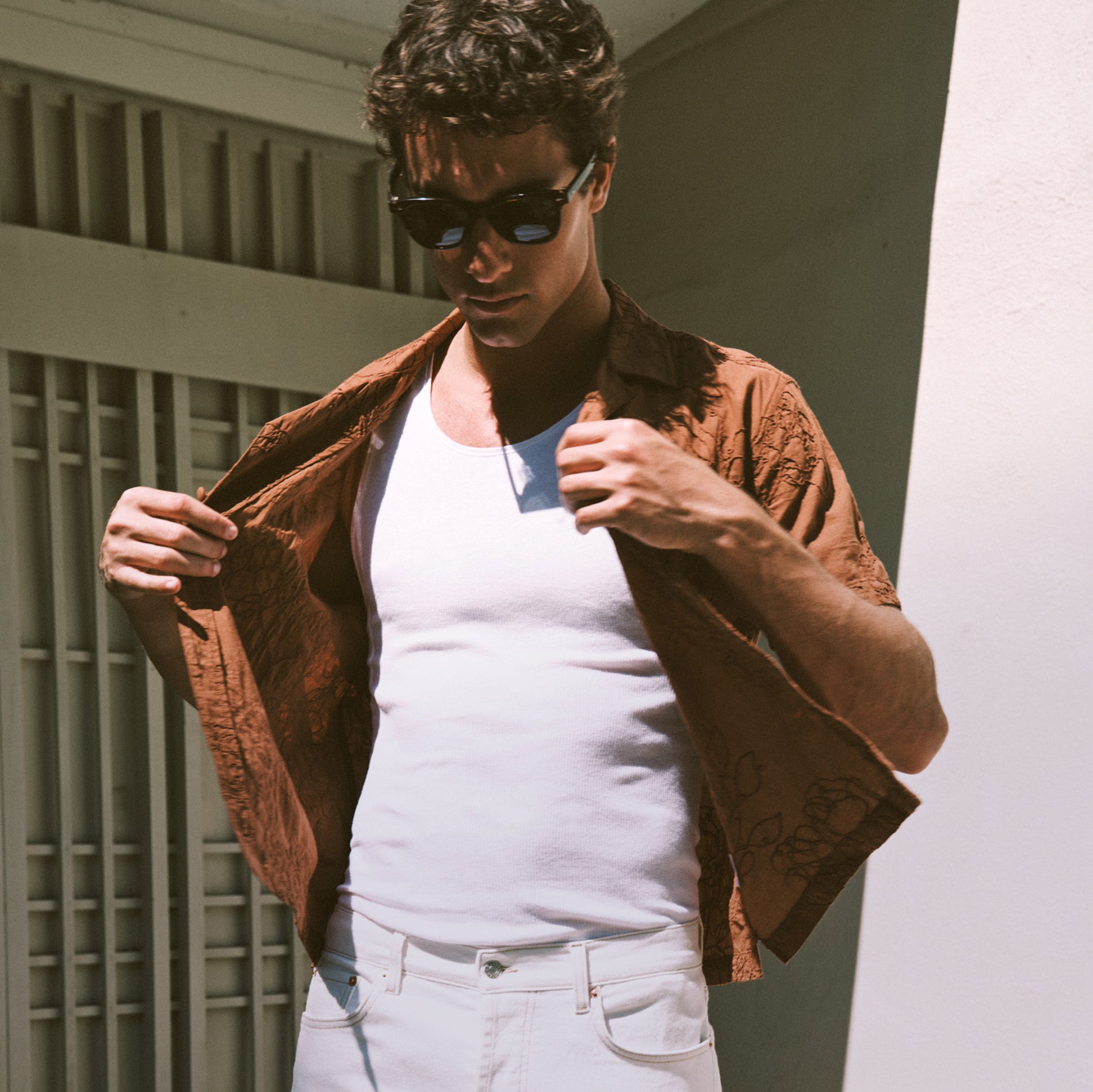 A man wearing black sunglasses, a white tank top, white jeans, and an open brown patterned short-sleeve button-up shirt, standing in front of a white wall and window slats.