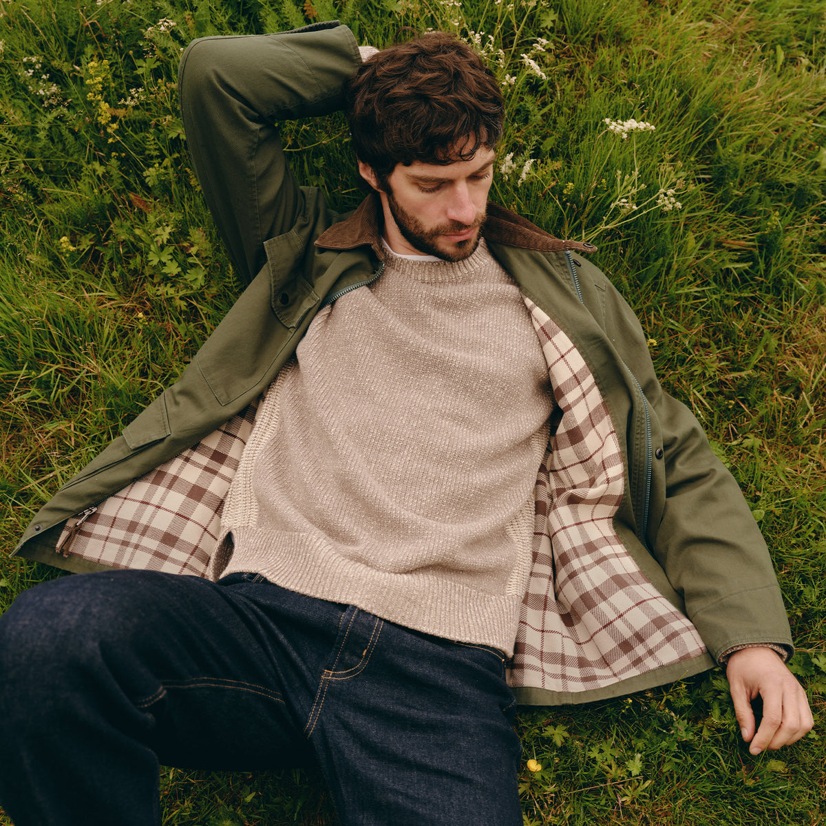 A man lying on green grass wearing an olive green jacket with a brown corduroy collar, a beige knit sweater, a brown and beige plaid shirt, and dark blue jeans.