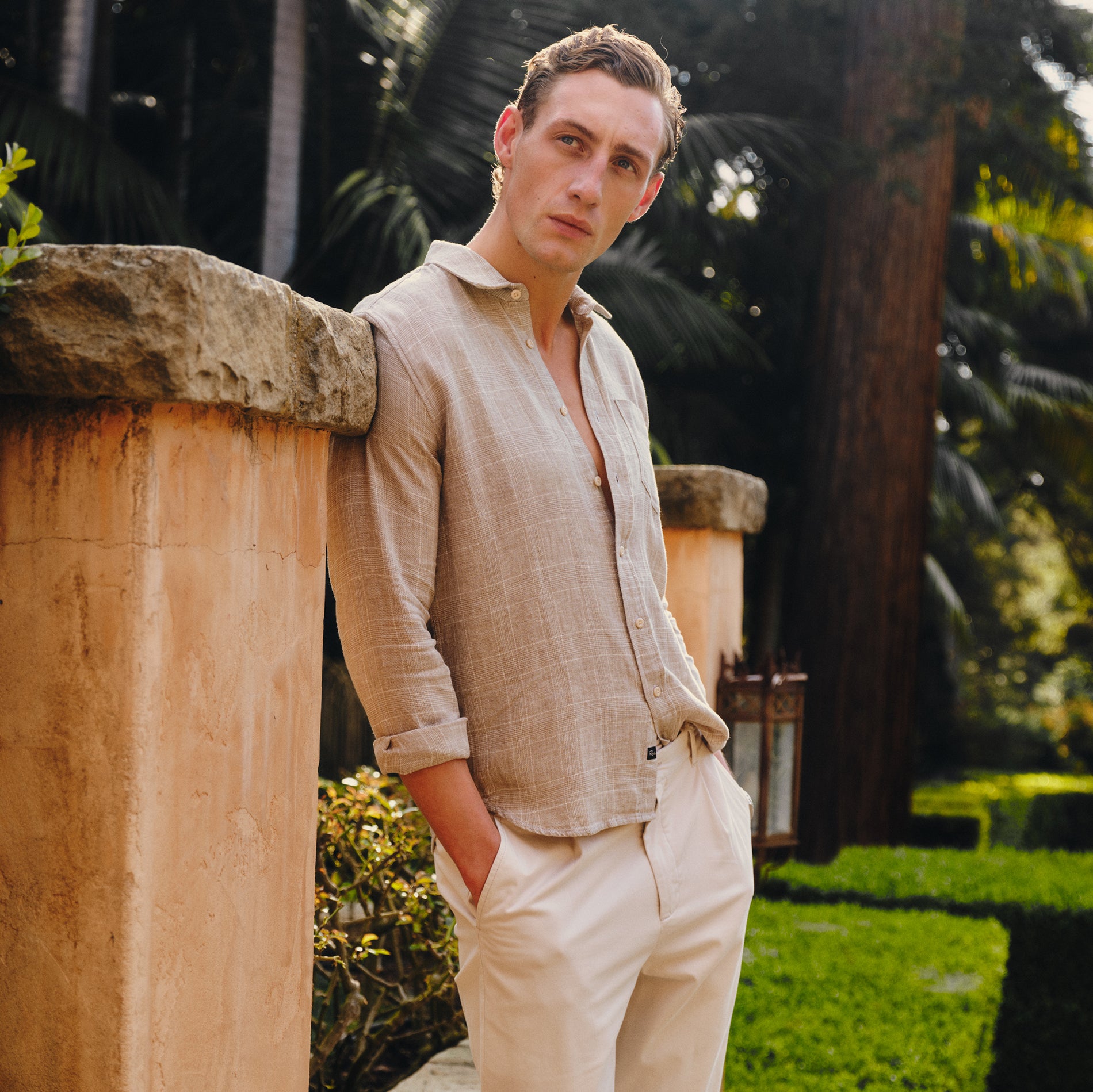 Man in beige shirt and pants leaning against a stone wall in a garden.