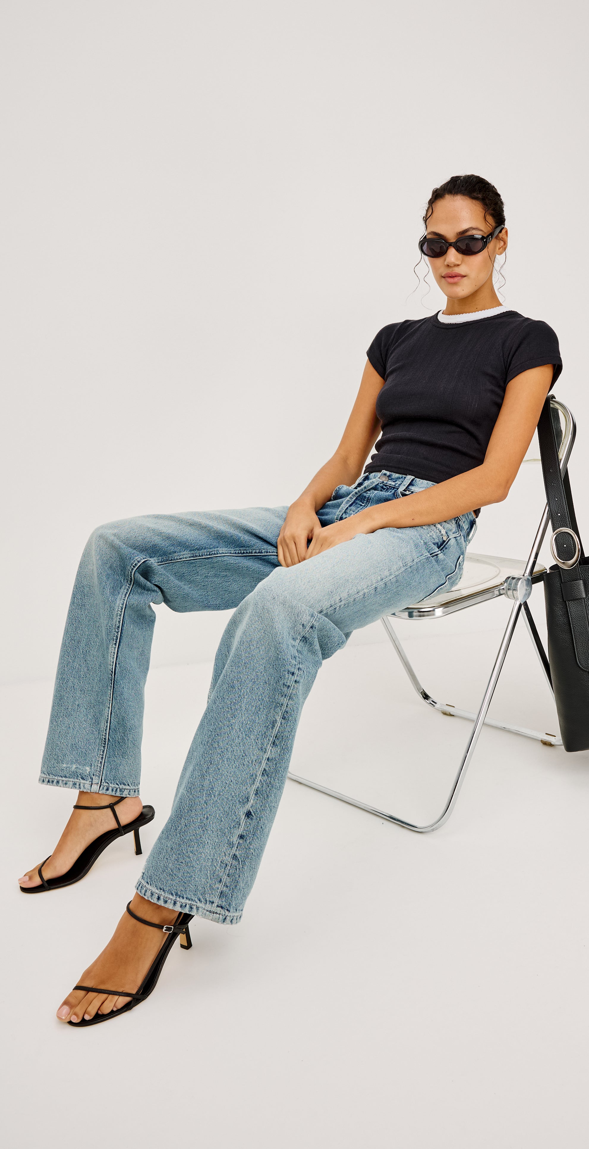 Woman sitting on a chair wearing a black top, blue jeans, and sunglasses with a black handbag beside her on a white background.