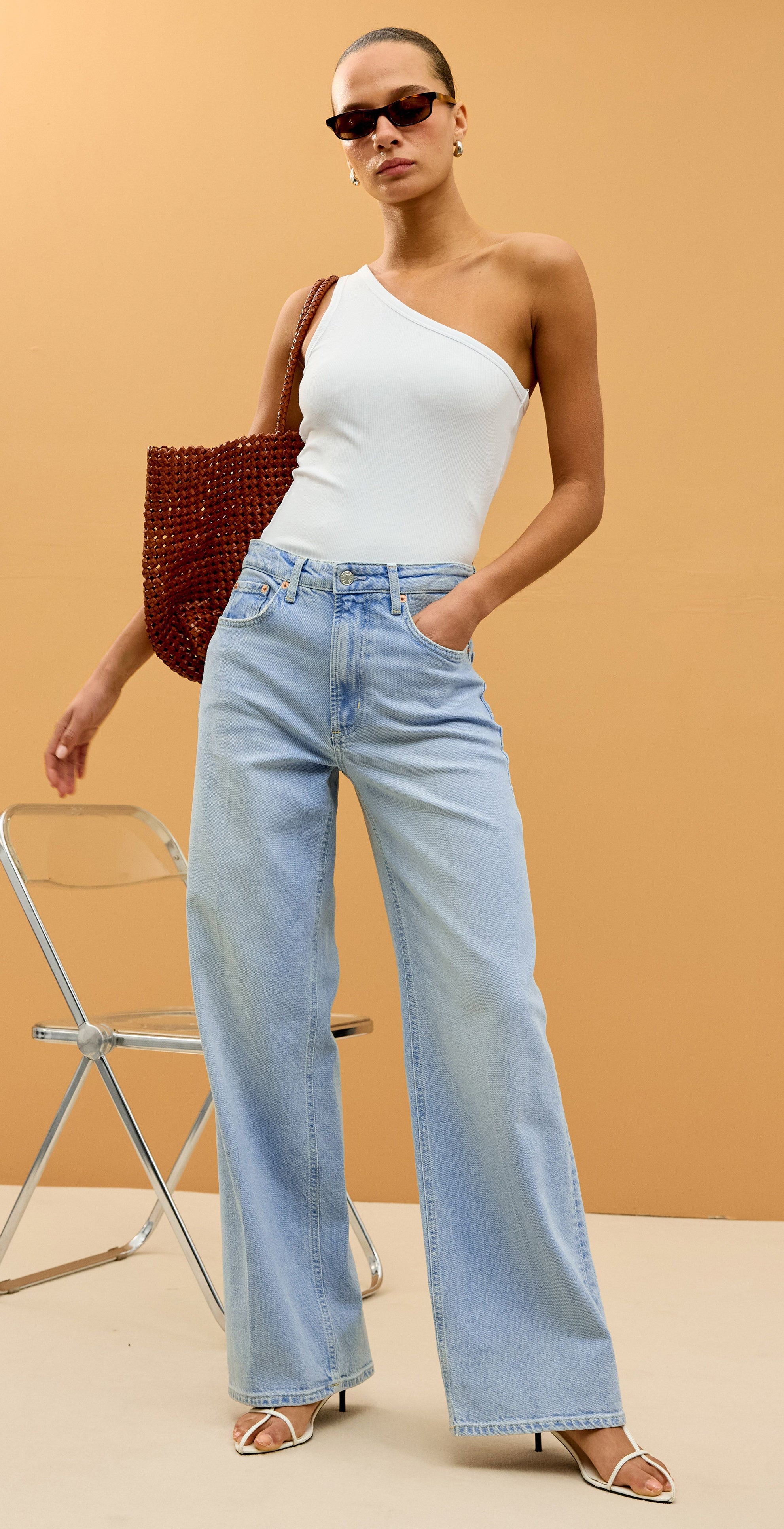 Woman wearing a white one-shoulder top and light blue jeans against an orange background