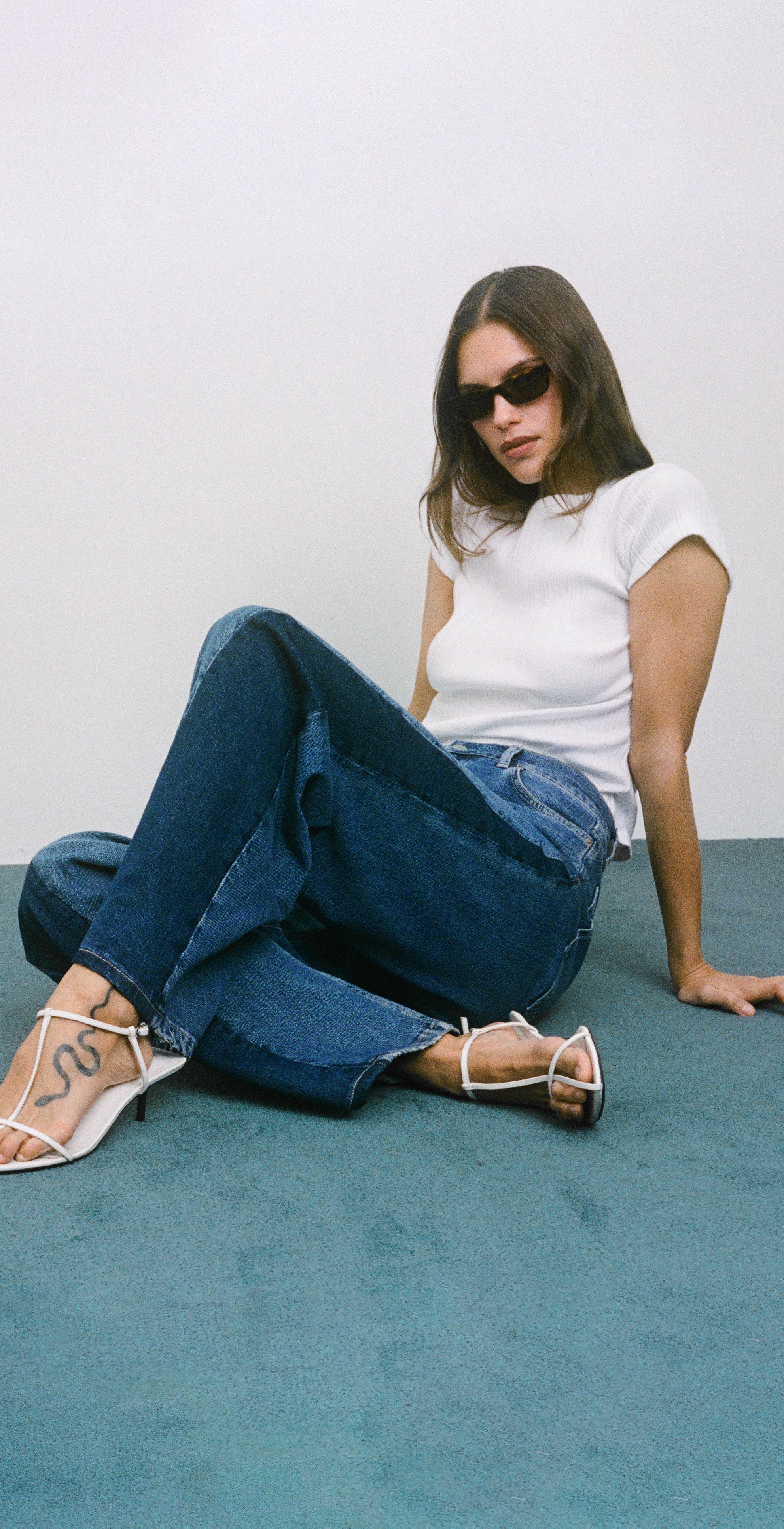 Woman sitting on a blue carpet wearing a white t-shirt, blue jeans, and white heels.