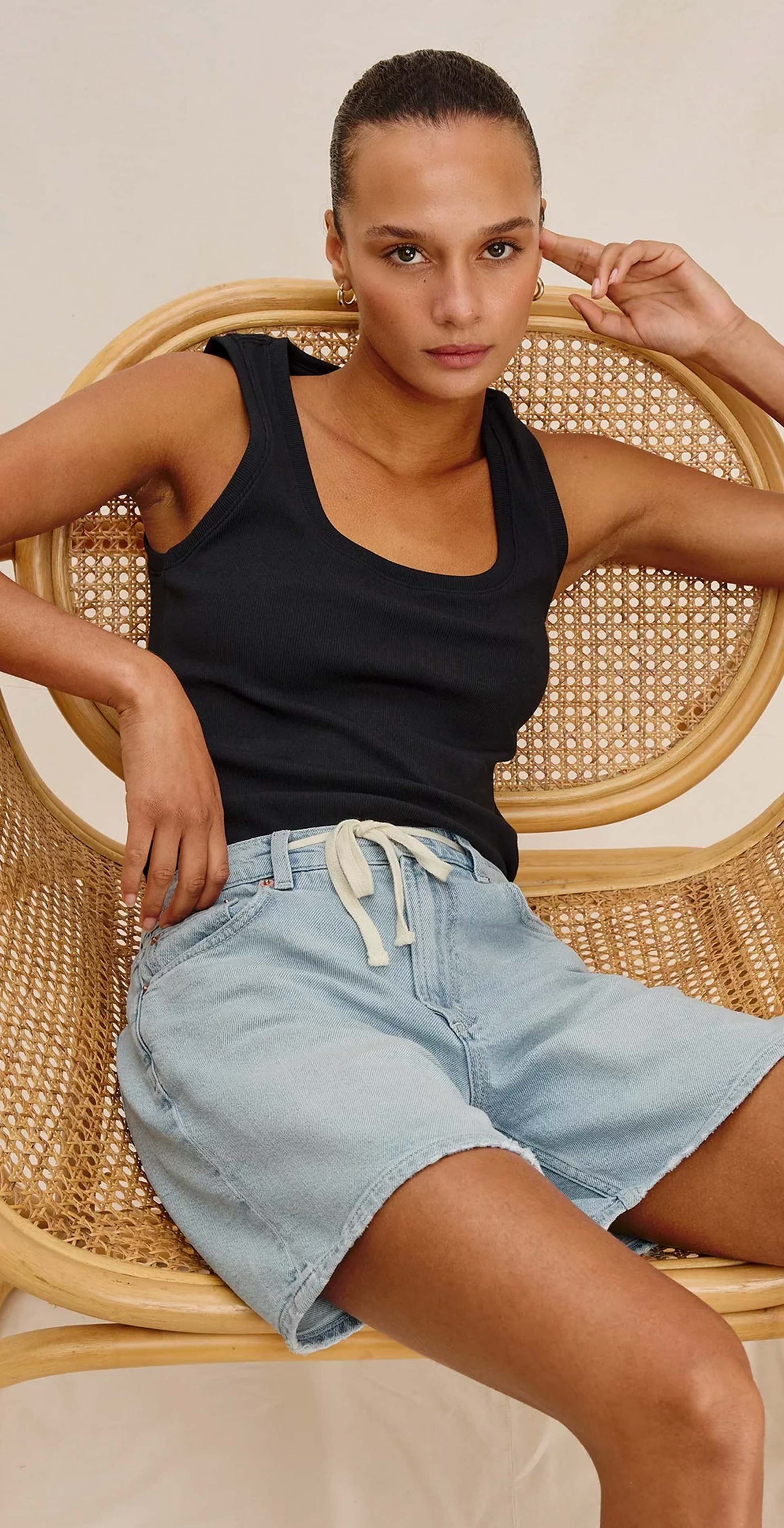 Woman sitting on a wicker chair wearing a black tank top and light blue shorts against a plain background