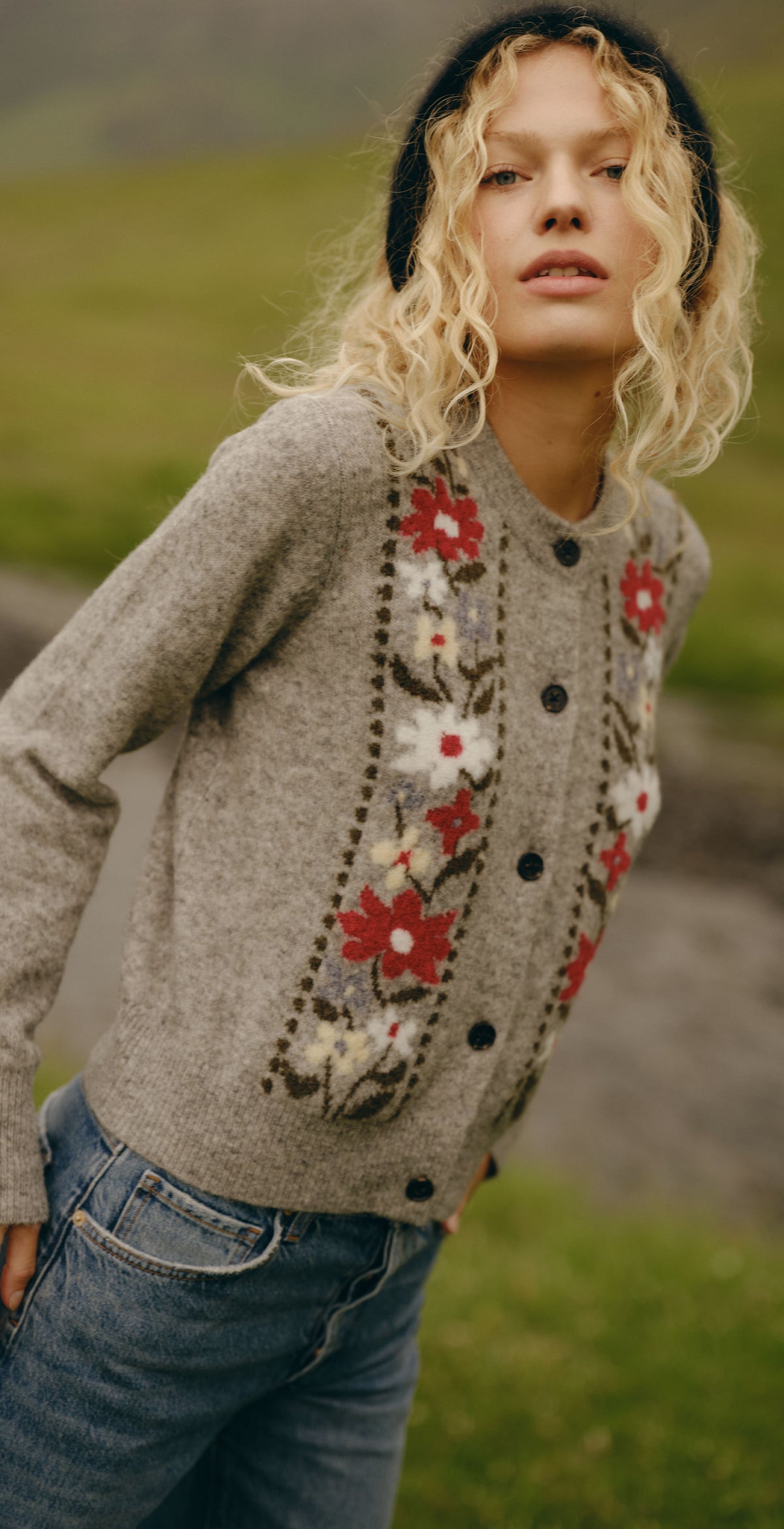 A split-screen lifestyle image showing a model wearing a gray knit cardigan with red and white floral embroidery, paired with blue jeans and black boots, in an outdoor grassy landscape.