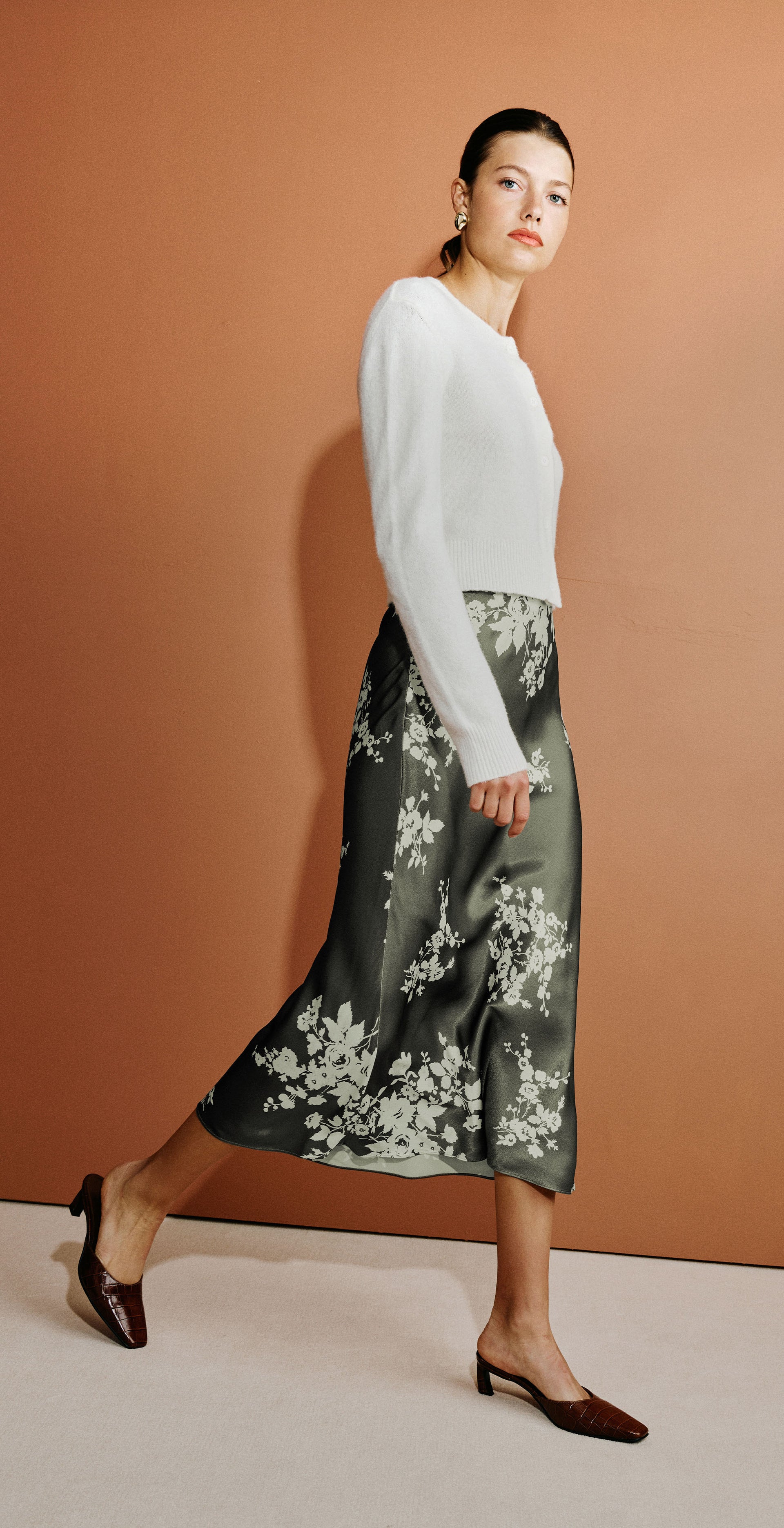 A model is shown in a split-screen fashion photo against a warm terracotta background. On the left, she sits on a dark chair wearing a white long-sleeve top and a dark green floral midi skirt with brown pointed-toe heels.