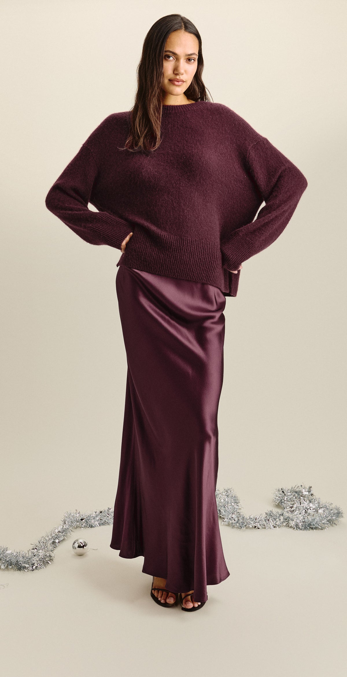 A woman modeling a two-piece outfit consisting of a fuzzy, oversized long-sleeve sweater and a satin-like floor-length skirt, both in a deep plum purple color, set against a plain beige background with silver tinsel on the floor.