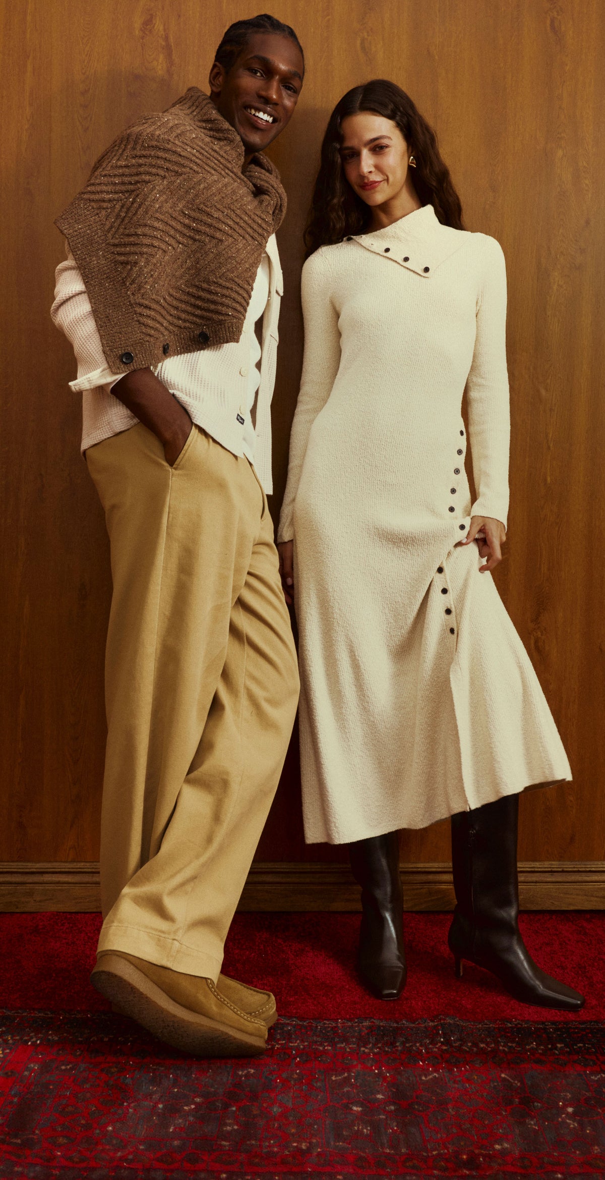Three models pose in a warm-toned indoor setting with wood-paneled walls and a red rug. The outfits feature cream knit turtleneck dresses, beige trousers, white shirts, and brown textured knit shawls.