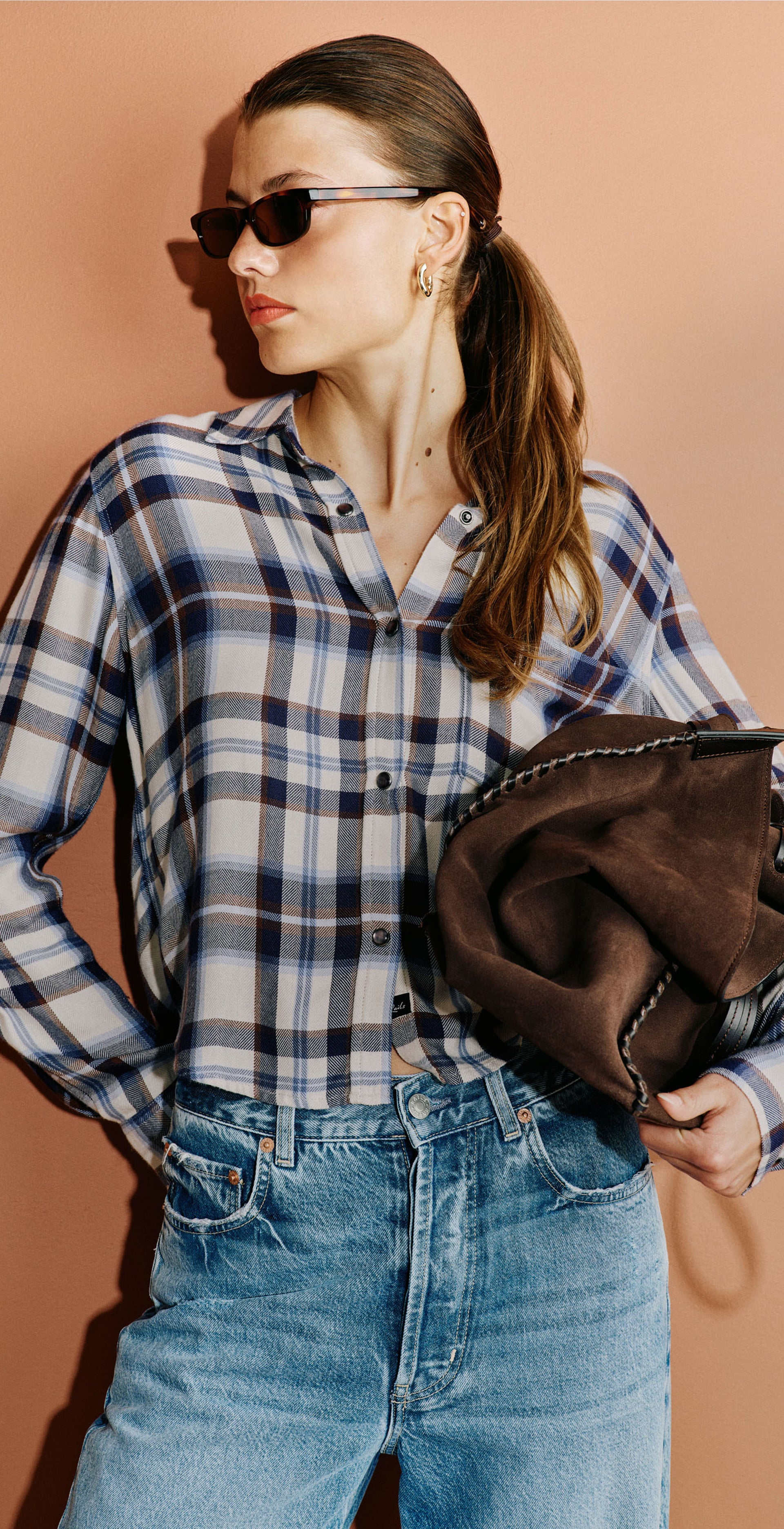A three-panel collage showing a woman modeling a blue, white, and black plaid button-down shirt with light-wash wide-leg jeans, sunglasses, and a dark brown shoulder bag against a solid terracotta background.