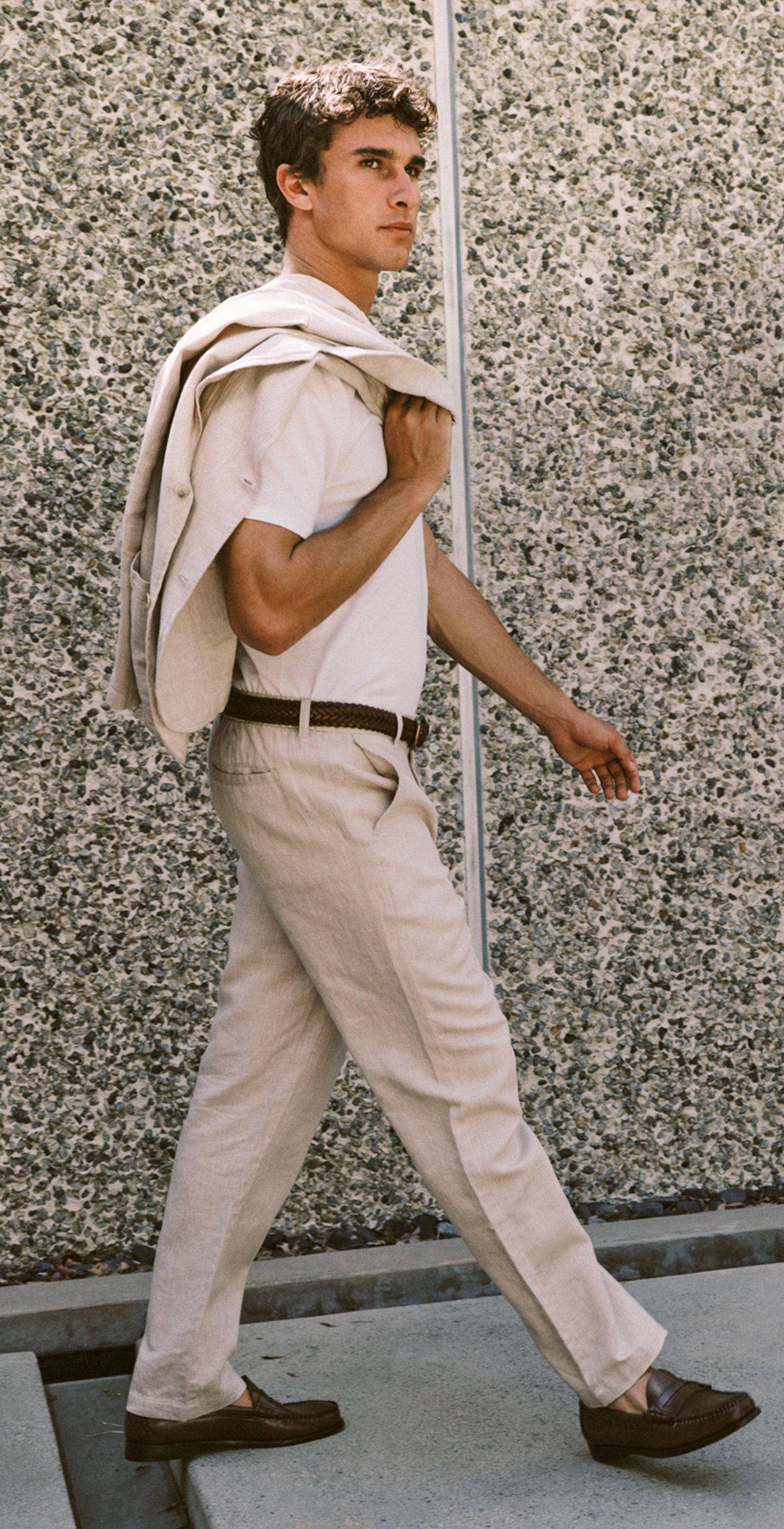 A man walking outdoors wearing a light beige short-sleeve shirt, matching beige trousers, and dark brown loafers, with a light-colored jacket draped over his shoulder, set against a textured concrete wall.