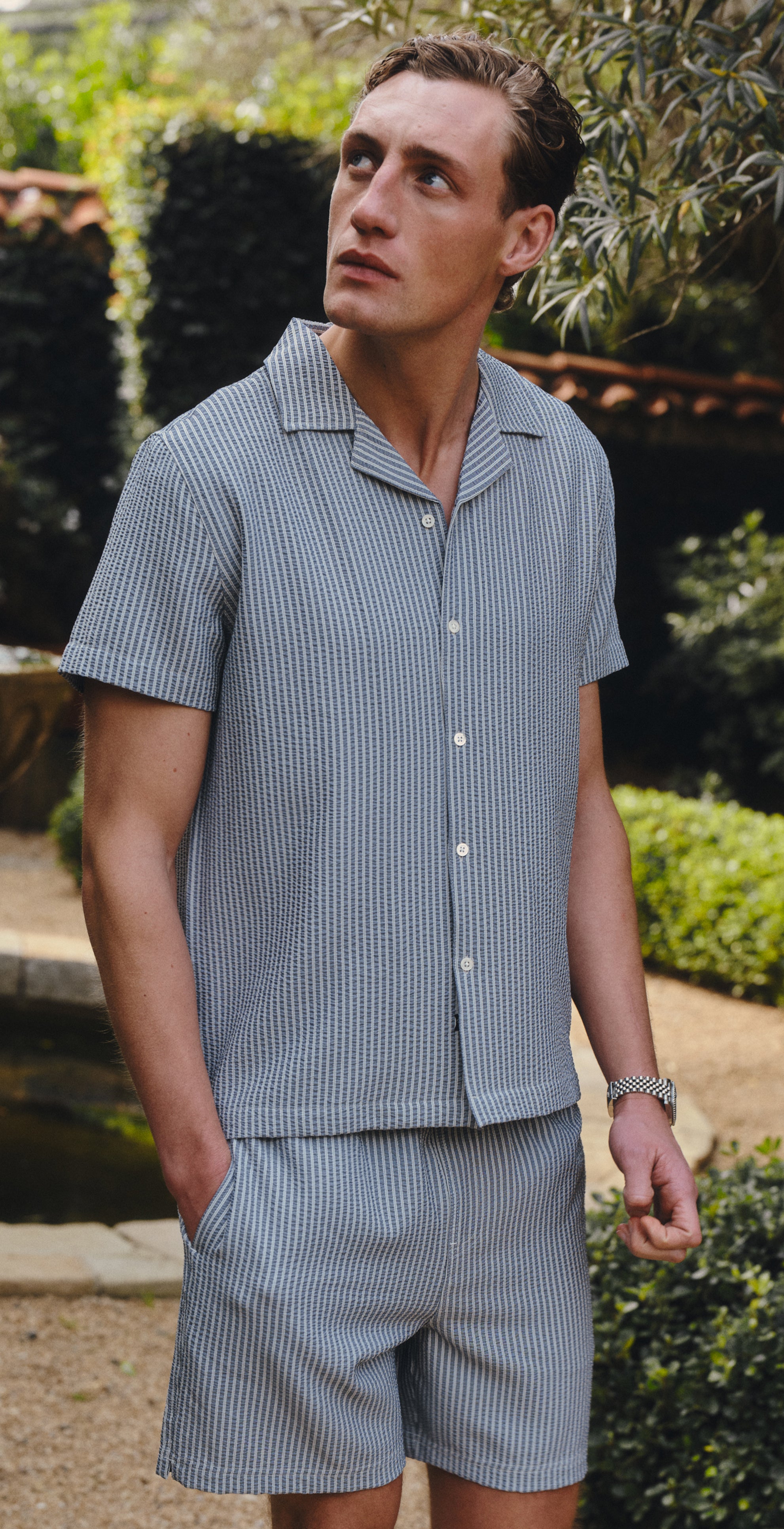 Man in a striped matching set standing in a garden with a fountain and greenery.