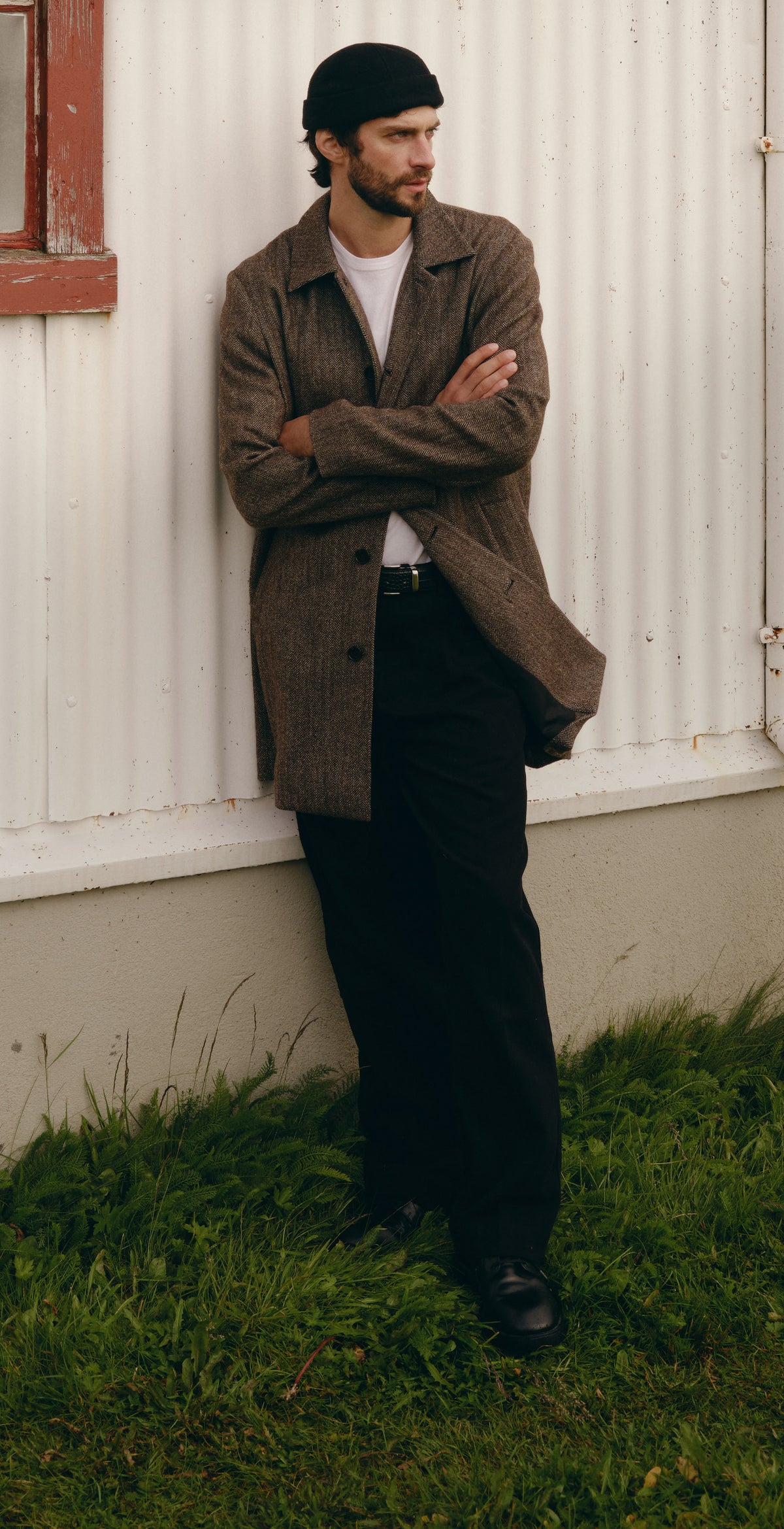 A man wearing a brown tweed jacket, black trousers, and a black beanie leans against the corner of a white house with a red-trimmed window, with grass and water in the background.