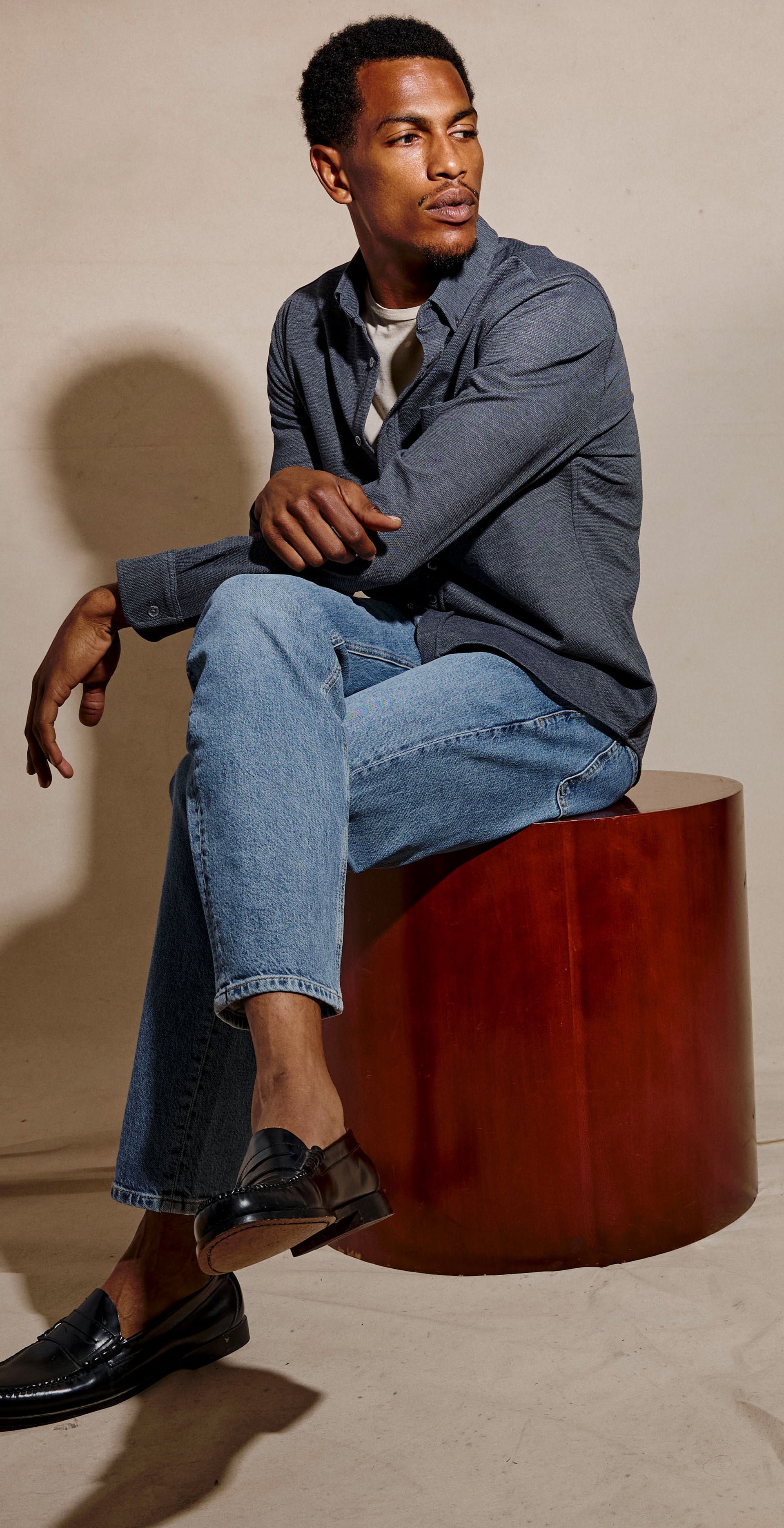 A man sits on a cylindrical wooden stool in a studio setting, wearing a gray blazer, light blue jeans, and black loafers against a warm beige backdrop.