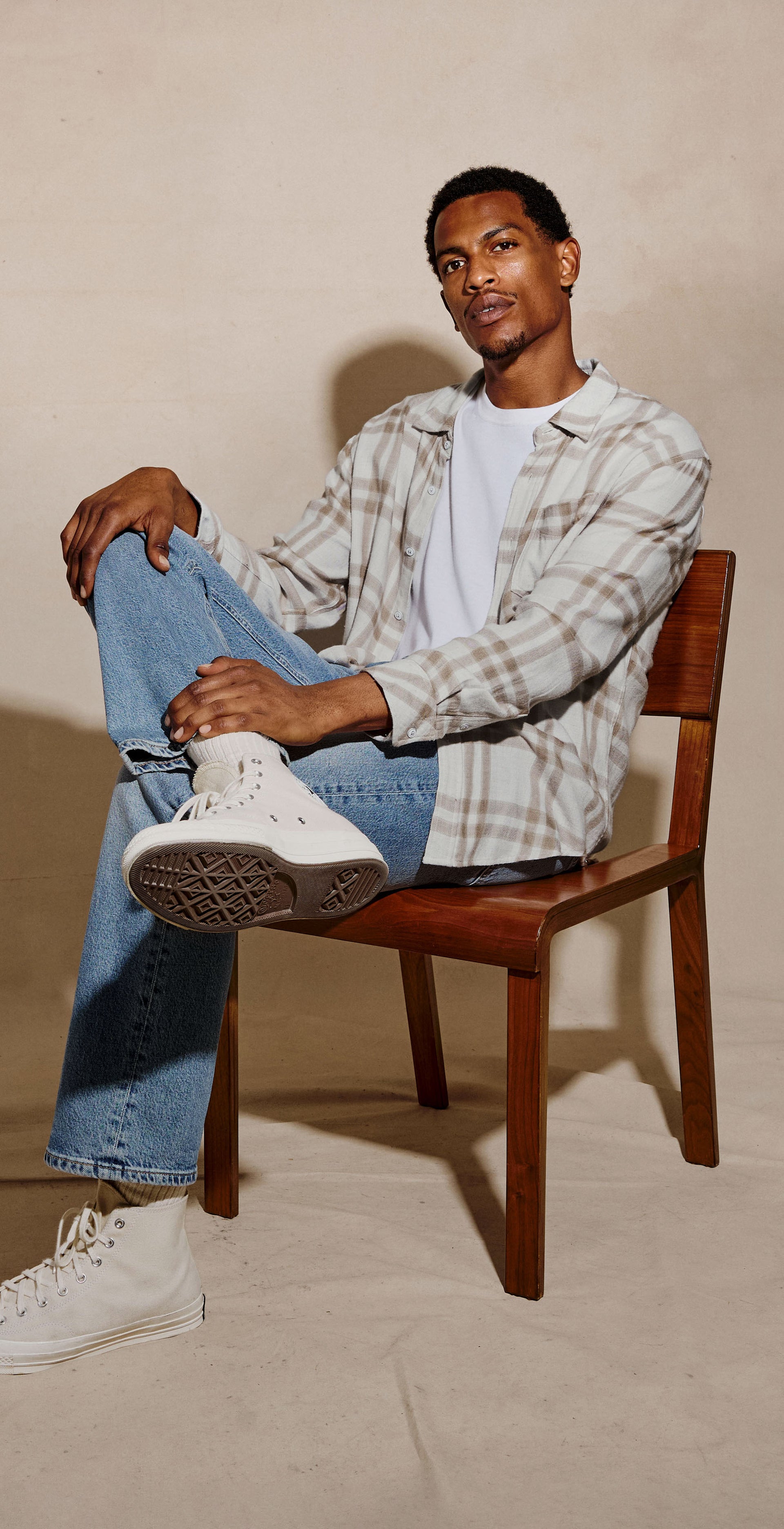 A man sits on a wooden chair against a warm beige studio backdrop, wearing a white and gray plaid button-up shirt over a white t-shirt, blue jeans, and white sneakers.
