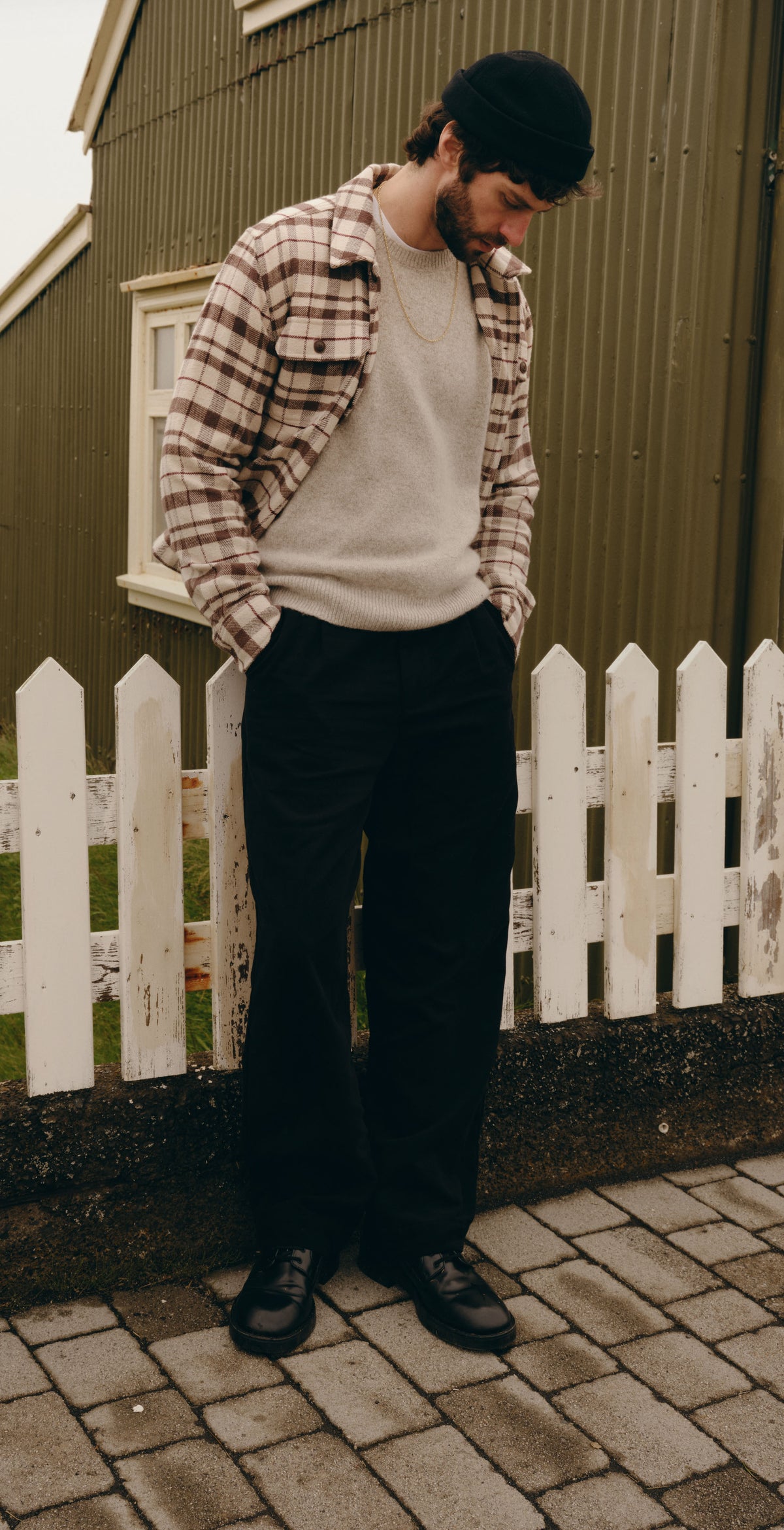 A man wearing a black beanie, a brown and white plaid flannel shirt over a white crewneck sweater, black trousers, and black shoes stands on a paved path next to a white picket fence and a dark green barn.