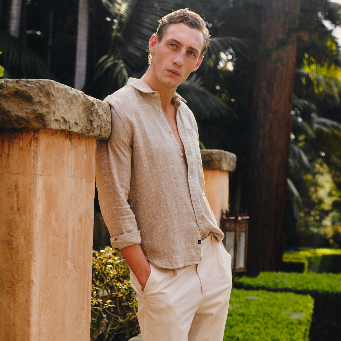 Man in beige shirt and pants standing outdoors with greenery in the background