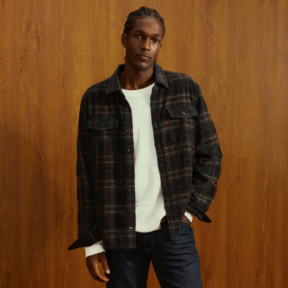 A man wearing a dark plaid button-up shirt over a white ribbed knit sweater and dark blue jeans, standing against a warm wood-paneled background.