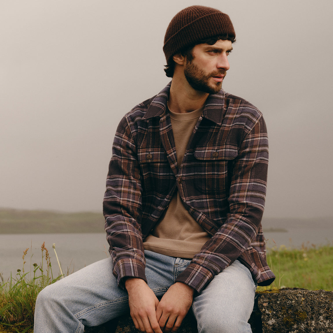 A man wearing a brown knit beanie, a brown and black plaid flannel shirt over a beige t-shirt, and light blue jeans, sitting outdoors on a stone wall with a misty lake in the background.