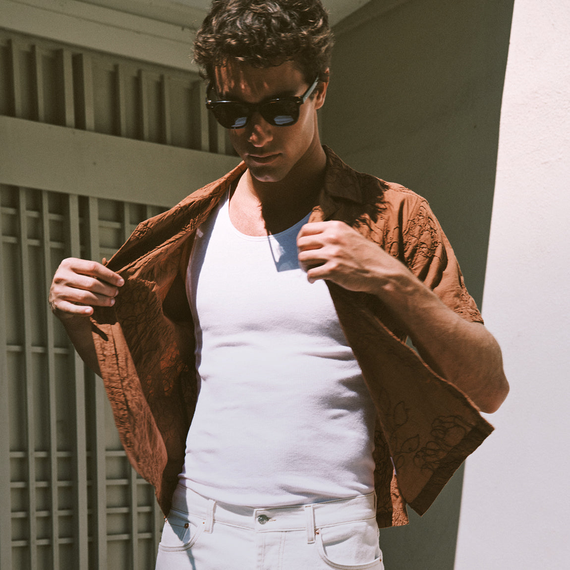 A man wearing black sunglasses, a white tank top, white jeans, and an open brown patterned short-sleeve button-up shirt, standing in front of a white wall and window slats.