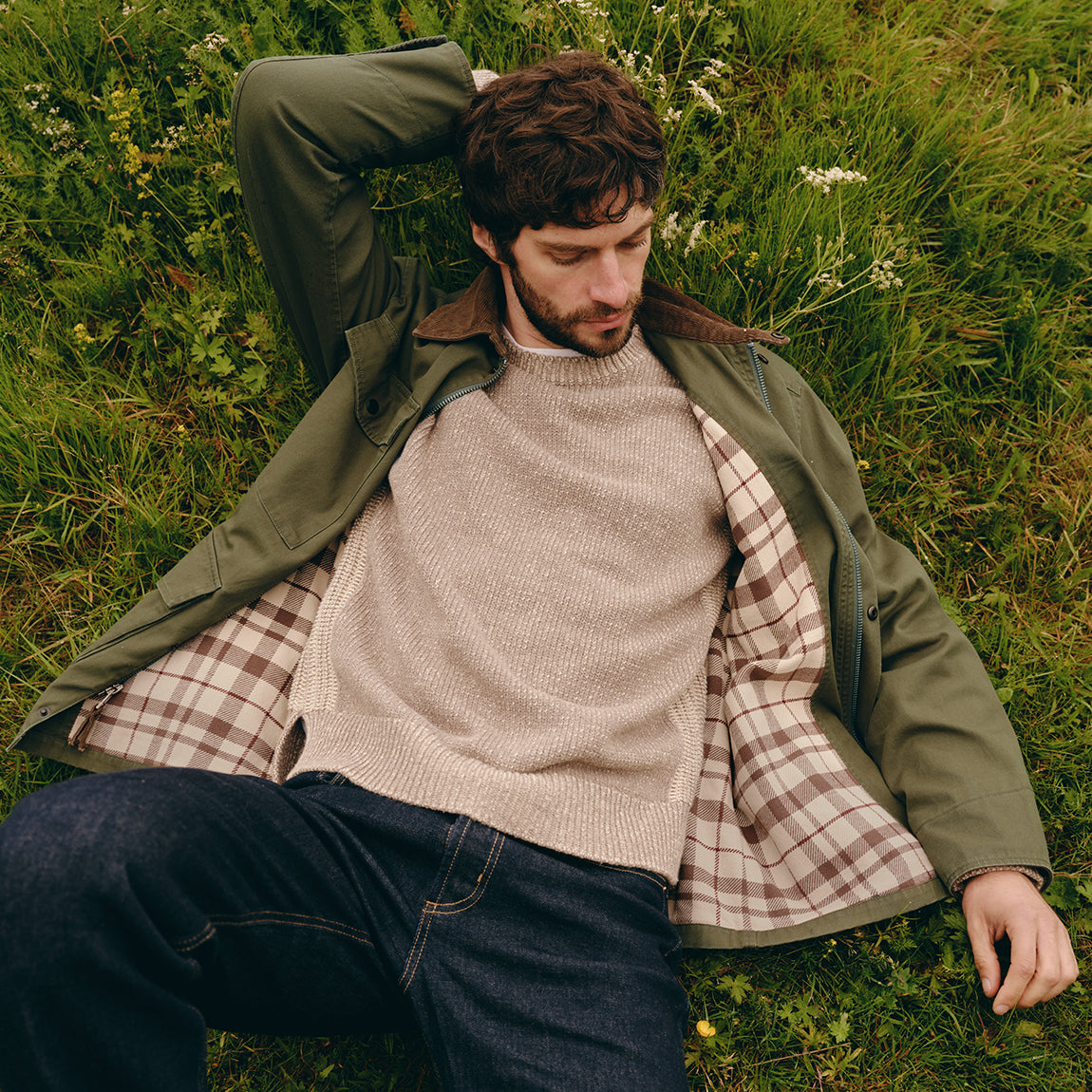 A man lying on grass outdoors wearing an olive green jacket over a beige knit sweater and a brown-and-white plaid shirt, with dark jeans.