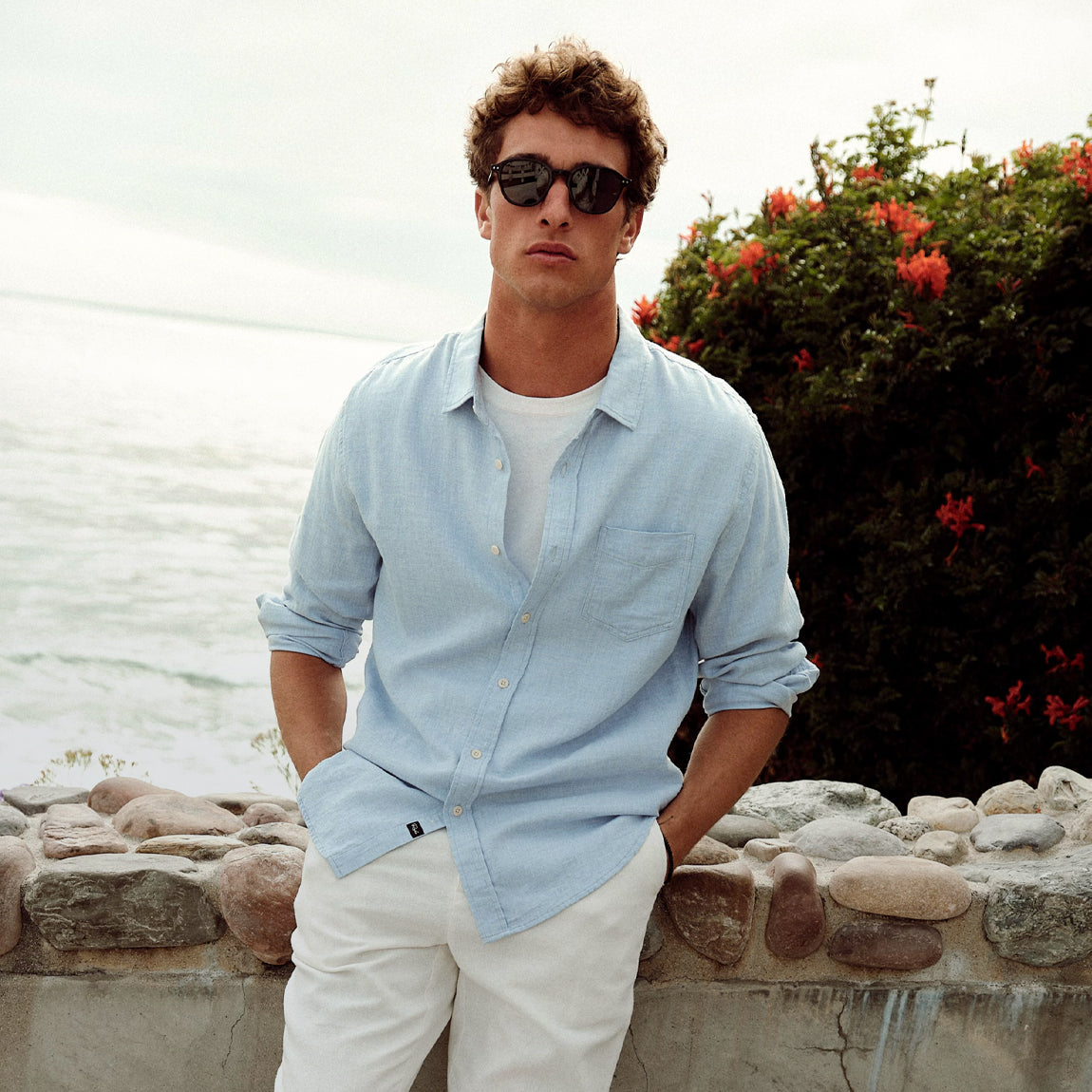 Man wearing sunglasses and a light blue shirt by a body of water with greenery.