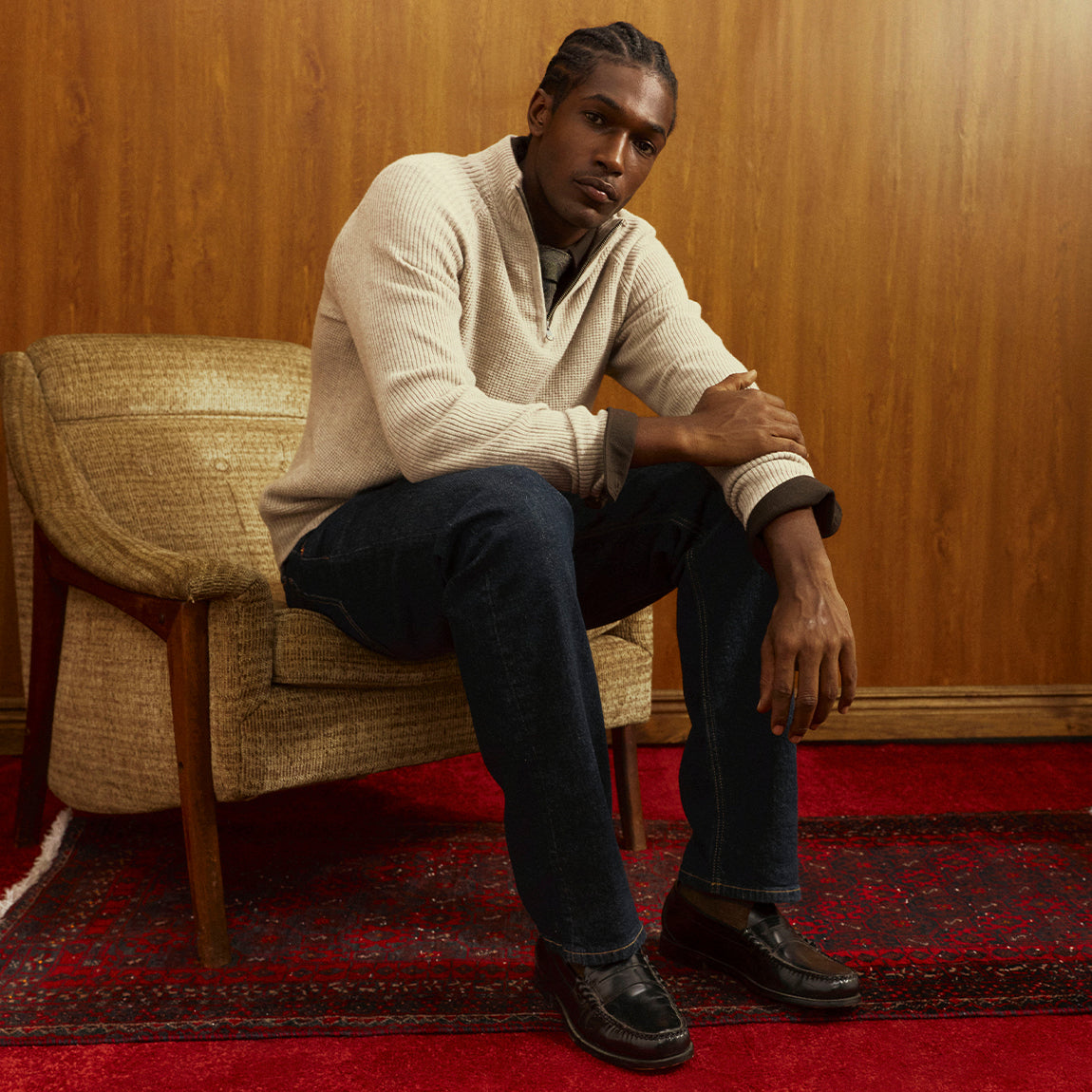 A man sits in a woven armchair in a wood-paneled room, wearing a cream ribbed knit quarter-zip sweater, dark blue jeans, and black leather loafers on a red patterned rug.