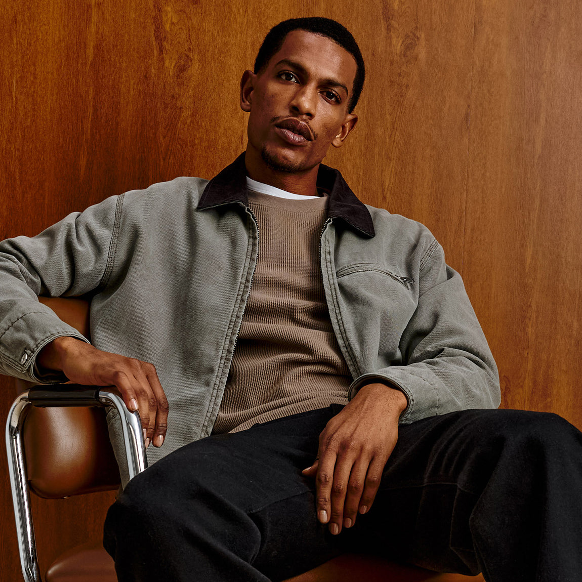 A man wearing a muted gray-green overshirt over a taupe knit sweater and black pants, seated on a brown leather chair against a warm wood-paneled wall.