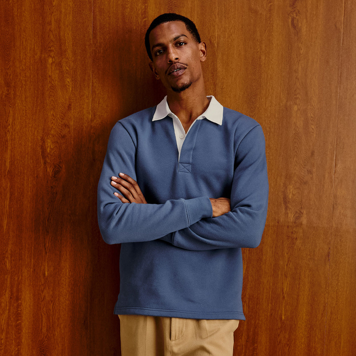 A man wearing a blue long-sleeve rugby shirt with a white collar, standing with arms crossed against a warm wood-paneled background.