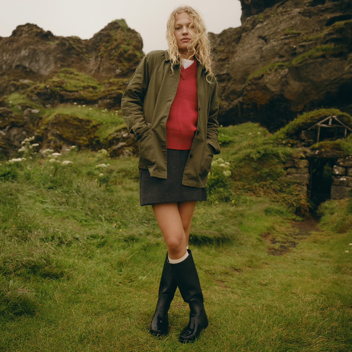 A woman stands outdoors in a grassy, rocky landscape wearing an olive green jacket, a red sweater, a dark gray skirt, white socks, and black rain boots.