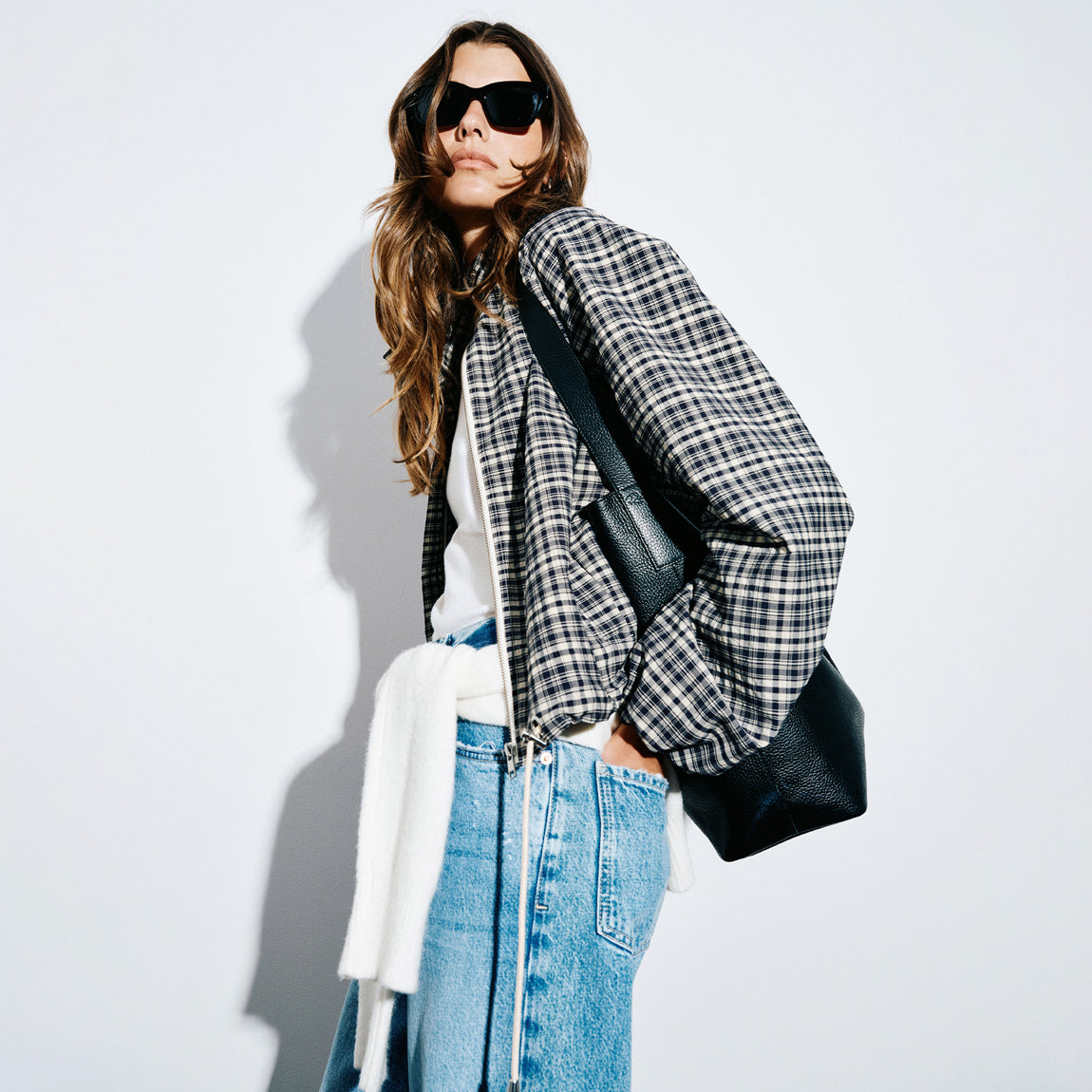 A woman models a black and white plaid puffer jacket, styled with a white knit sweater draped off her shoulder, blue denim skirt, and black sunglasses, carrying a black leather shoulder bag against a light gray background.