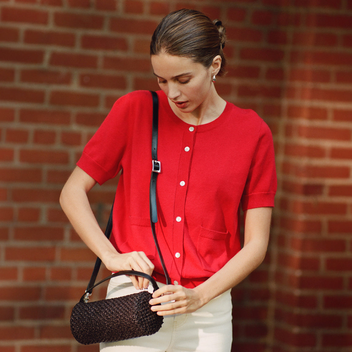 Woman in a red shirt holding a black woven handbag against a brick wall.