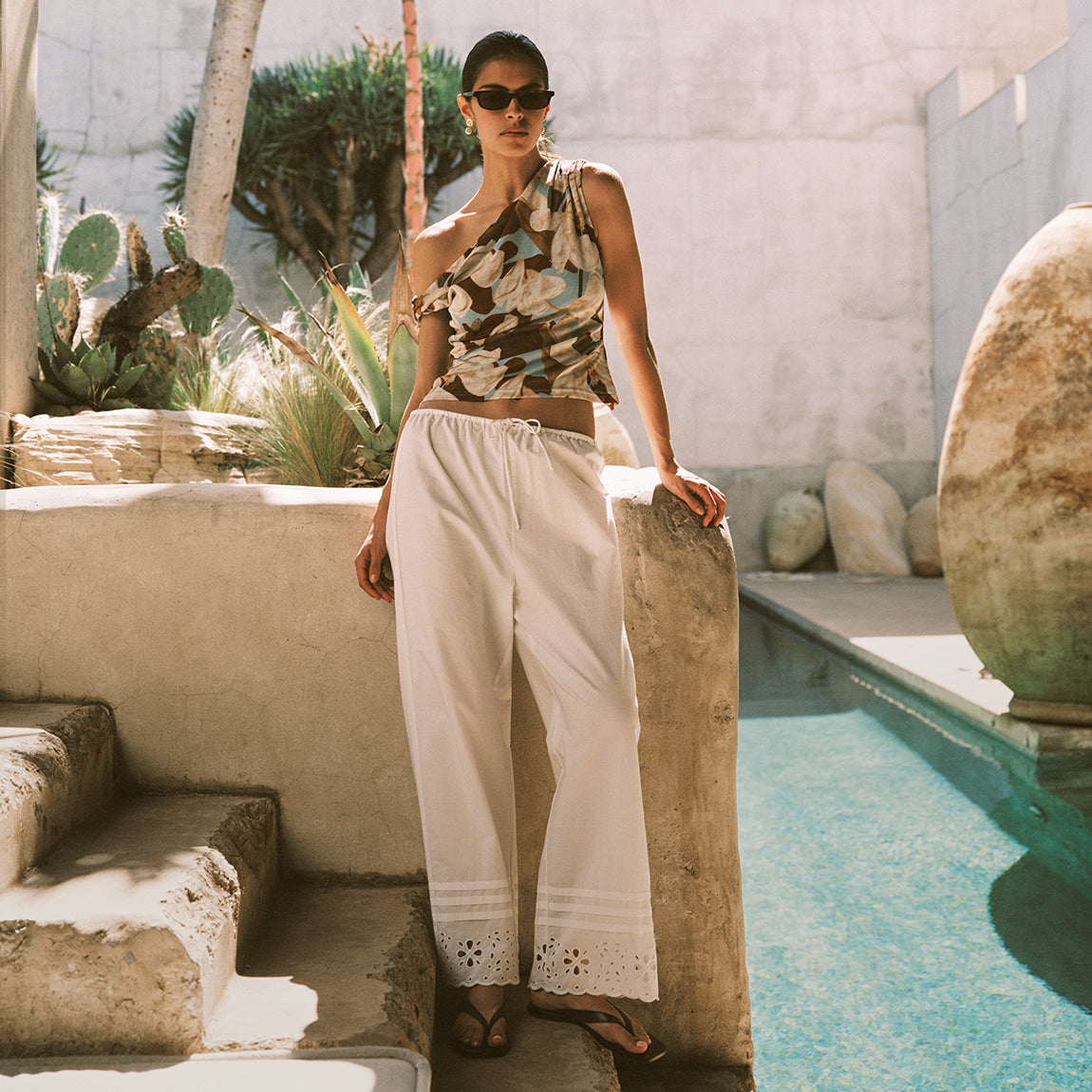 A woman wearing a sleeveless floral print halter top and white wide-leg drawstring pants stands beside a swimming pool in a sunlit outdoor setting with stone walls and plants.