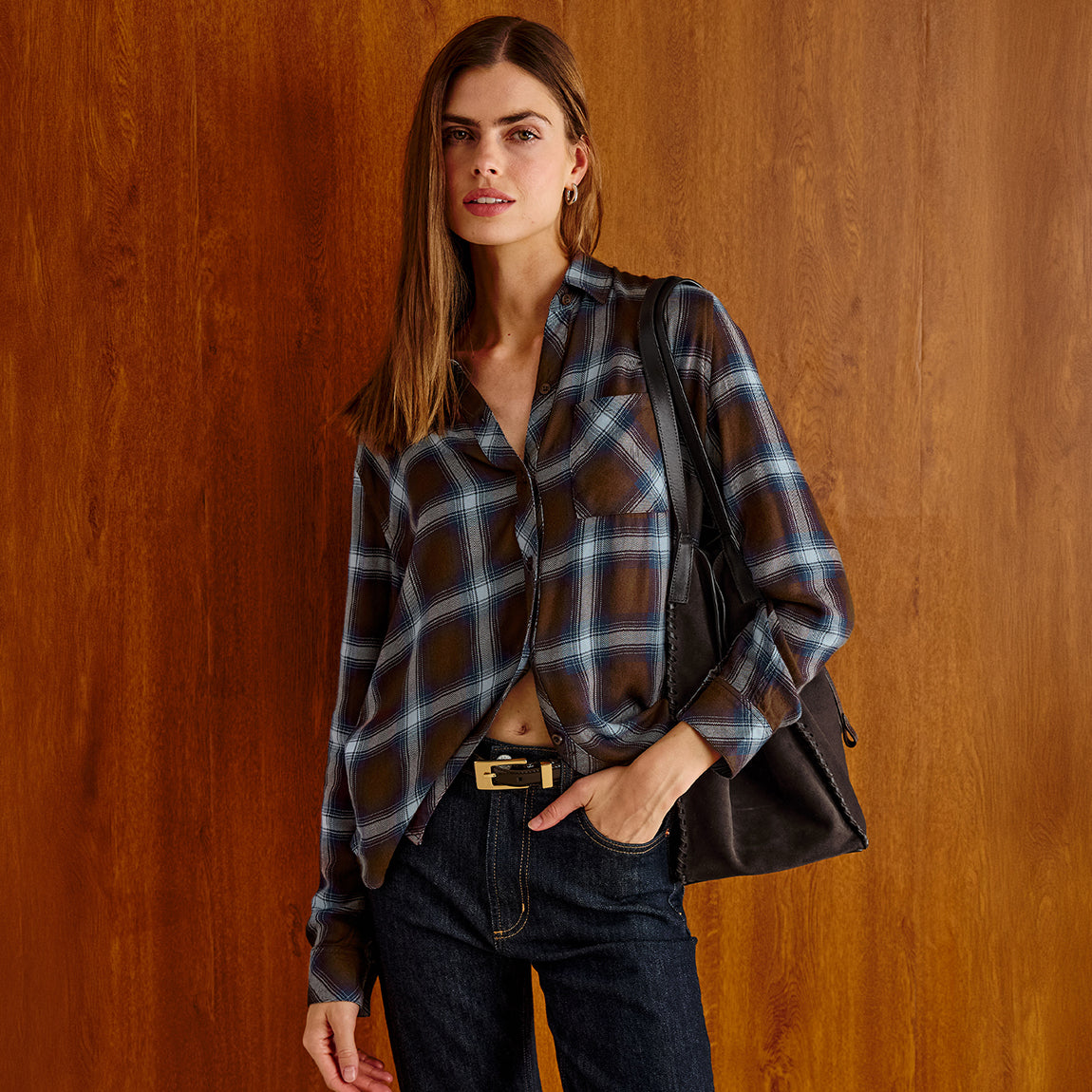 A woman models a brown and blue plaid button-down shirt paired with dark jeans and a black shoulder bag, standing against a warm wood-paneled wall.