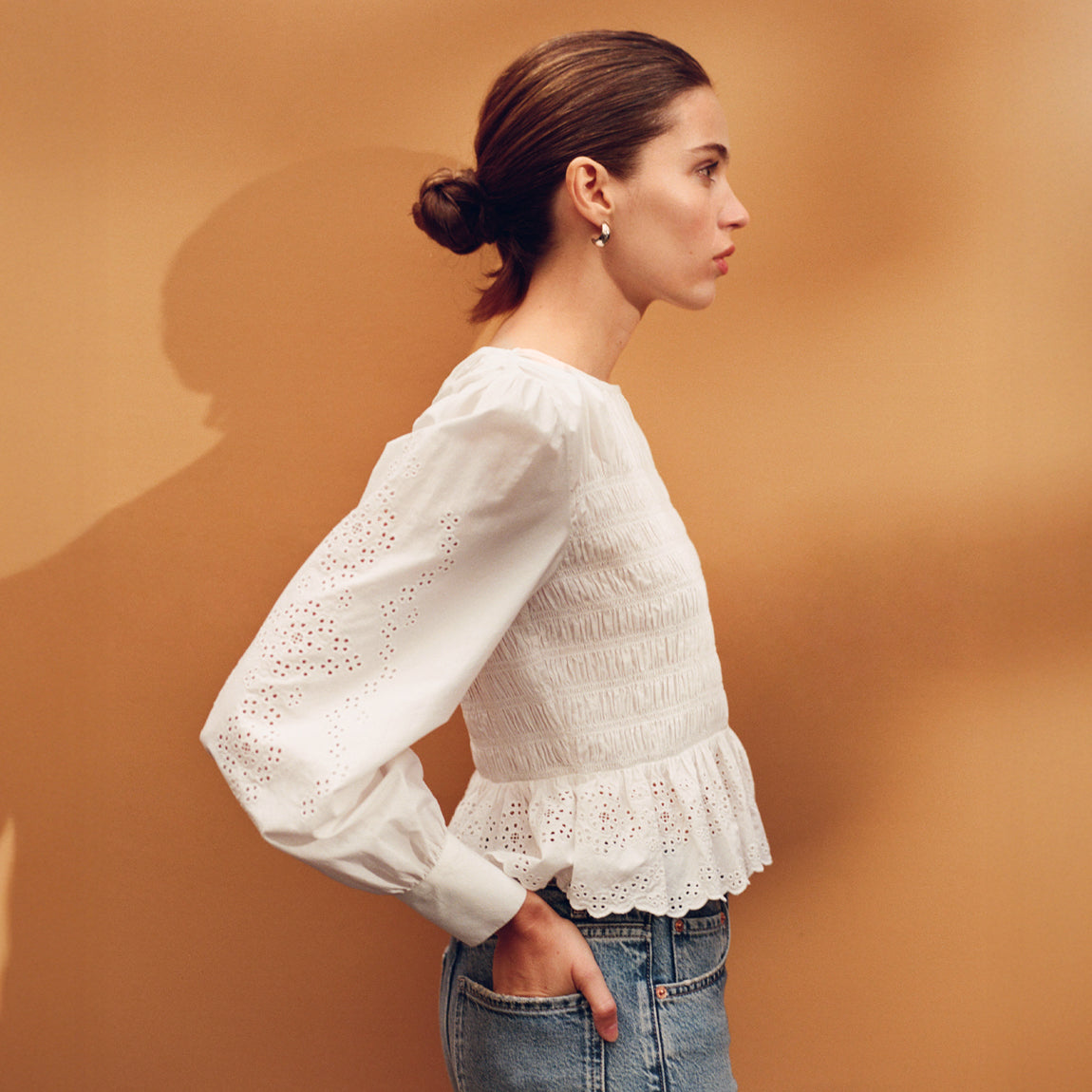 Woman wearing a white blouse with long sleeves and a ruffled bottom against a brown background.