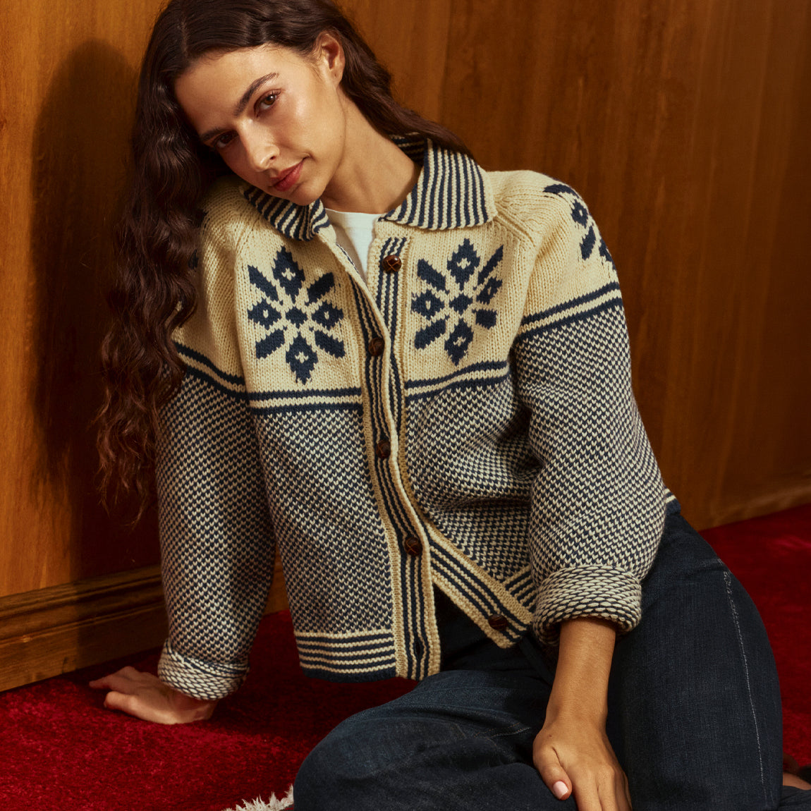 A woman wearing a cream and navy patterned knit cardigan with a button front, sitting on a red rug against a wooden wall.