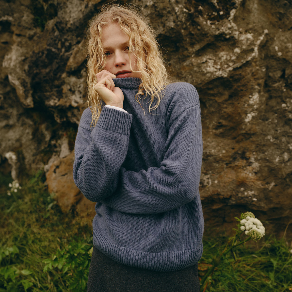 A woman with curly blonde hair wearing a slate blue ribbed turtleneck sweater, standing outdoors in front of a large rock face with green grass and small white flowers at her side.