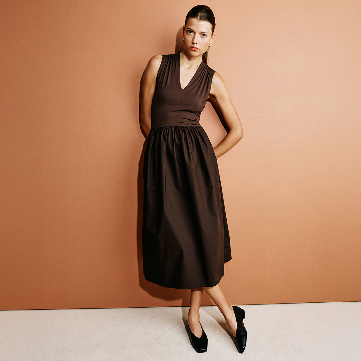A woman models a sleeveless dark brown midi dress with a V-neckline and gathered waist, paired with black flats, set against a solid terracotta background.