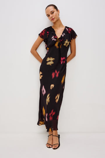 A chiffon midi dress with a blurred floral print. The dress has a V-neckline framed by cascading ruffles and a velvet bow, ruffle cap sleeves, and an empire waist that creates a graceful, elongated silhouette. Full Body Front View featured on Model.