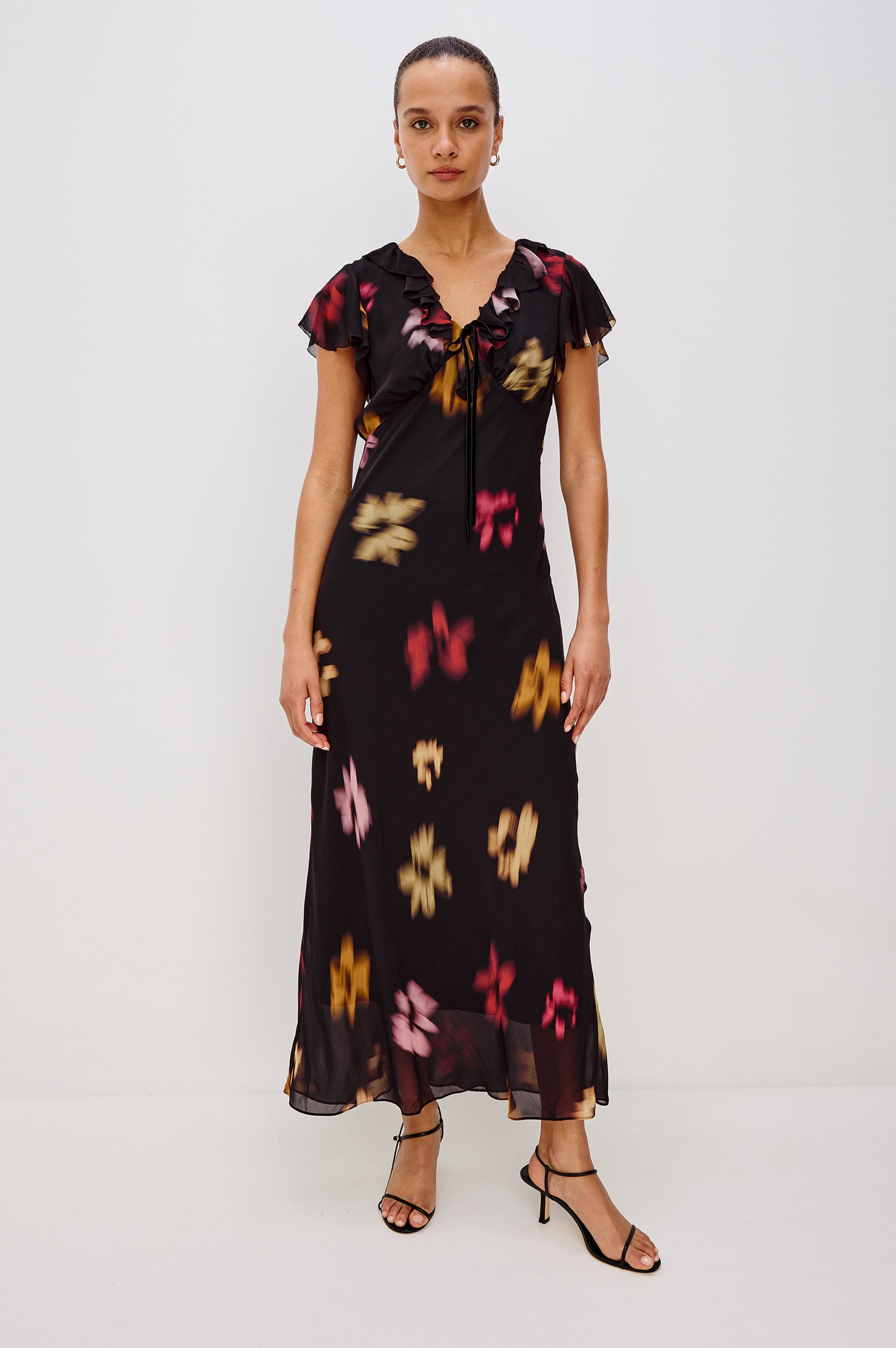 A chiffon midi dress with a blurred floral print. The dress has a V-neckline framed by cascading ruffles and a velvet bow, ruffle cap sleeves, and an empire waist that creates a graceful, elongated silhouette. Full Body Front View featured on Model.