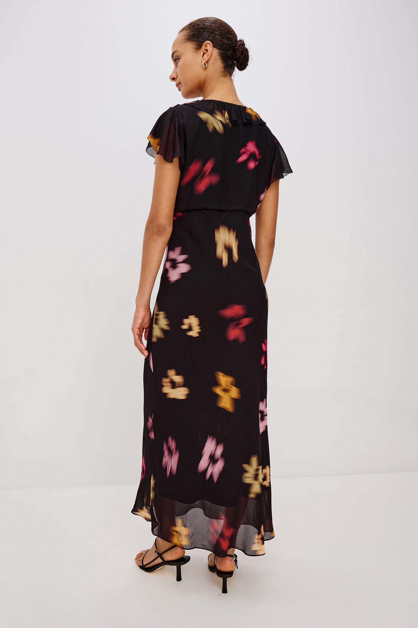 A chiffon midi dress with a blurred floral print. The dress has a V-neckline framed by cascading ruffles and a velvet bow, ruffle cap sleeves, and an empire waist that creates a graceful, elongated silhouette. Full Body Back View featured on Model.