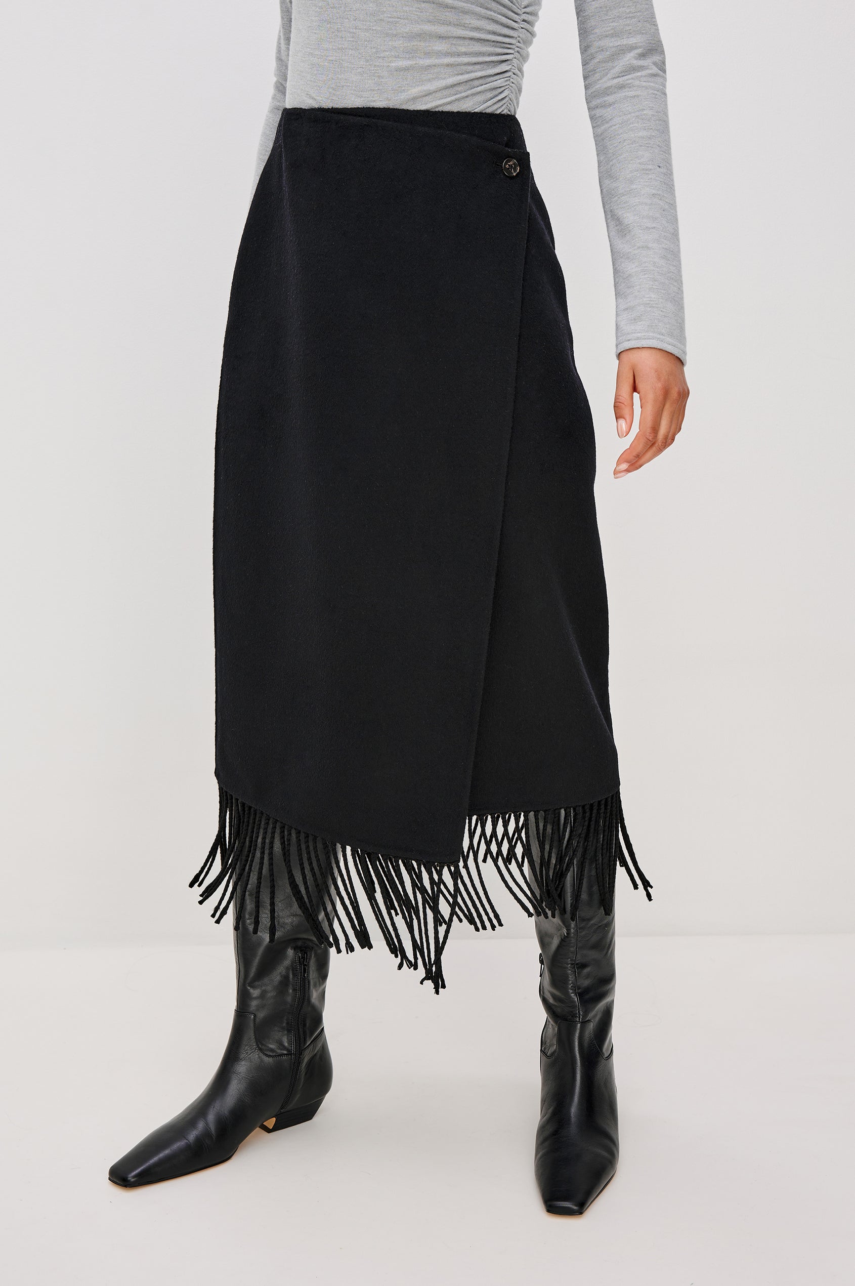 A high-waisted black wool-blend midi skirt. The wrap-style design fastens with a side button and interior closure, finished with statement fringe along the hem for added movement. Front View Featured on Model.