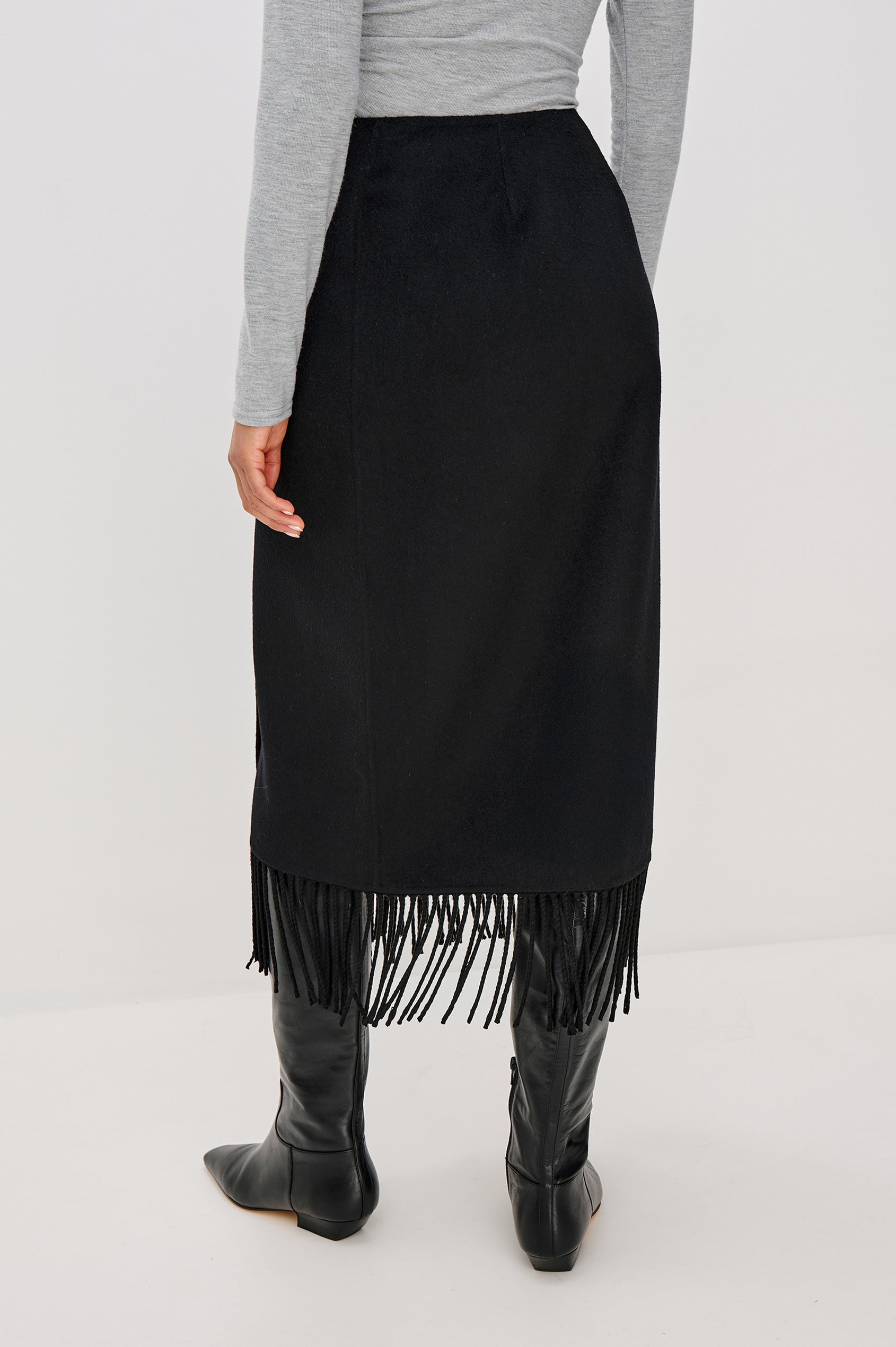A high-waisted black wool-blend midi skirt. The wrap-style design fastens with a side button and interior closure, finished with statement fringe along the hem for added movement. Back View Featured on Model.