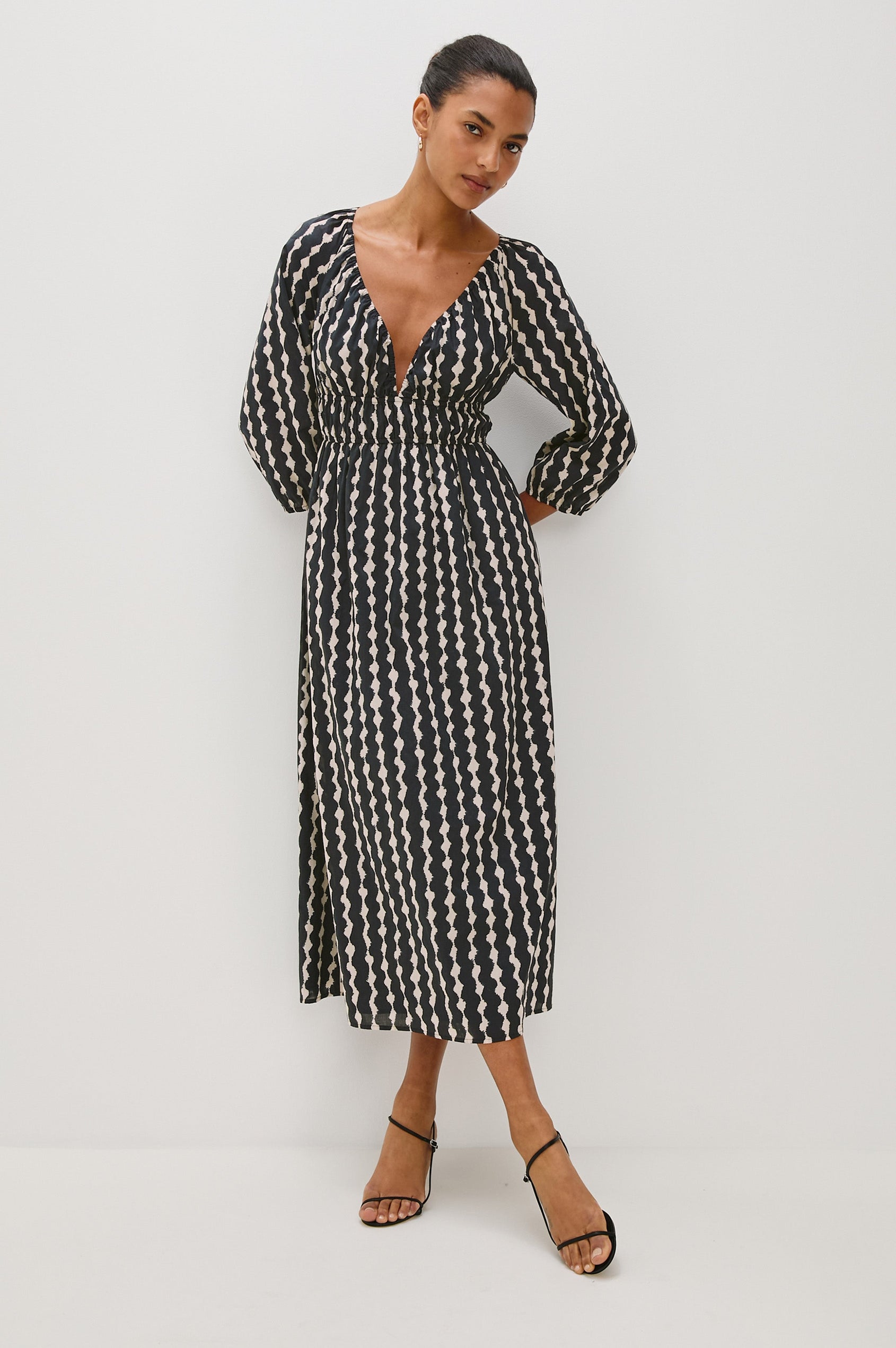 Paio midi dress with a fitted bodice, three quarter sleeves with a slight puff effect, flowing skirt, and eyelet closures at the chest in Painted Noir colorway - full body front view of model posing with hands behind back