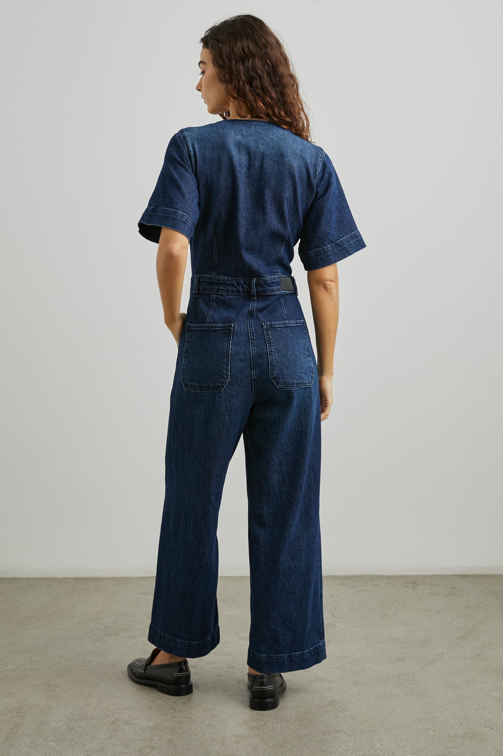 Palisades Short Sleeve Denim Jumpsuit with Wide Legs and Sleeves, Belt Loops, A Button and Zipper Closure for the Pants and Button Closure on the Bodice, a V neck, two Front Pockets and Two Back Patch Pockets in Blue Moon Colorway - Back Full Body View Featured on Model