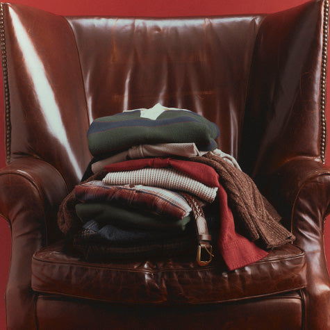 A stack of folded knit and woven garments in muted earthy tones resting on the arm of a dark brown leather armchair against a solid red background.