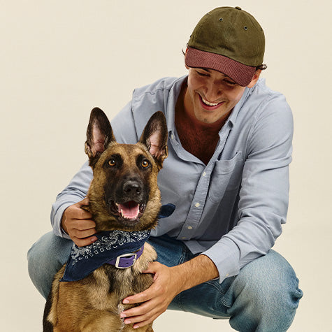 A smiling man crouches beside a German Shepherd dog wearing a navy blue bandana with a purple buckle. The man wears a light blue button-up shirt, blue jeans, and an olive green baseball cap.