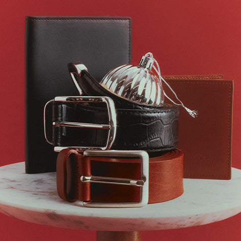 Three leather belts in black, brown, and reddish-brown are arranged on a white marble pedestal against a solid red background, with a black wallet, brown cardholder, and a silver decorative dome behind them.