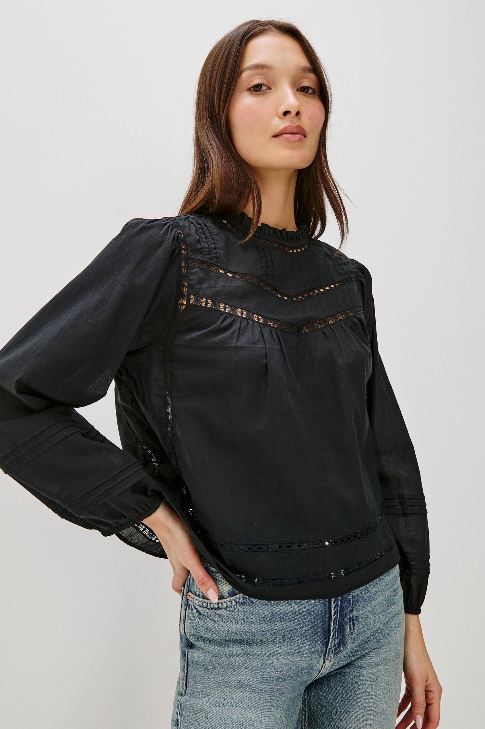 Front view of female model wearing our Reyna Top in black. This is a long sleeve crewneck baby doll blouse, with ruffles at the collar. Styled with light wash denim.