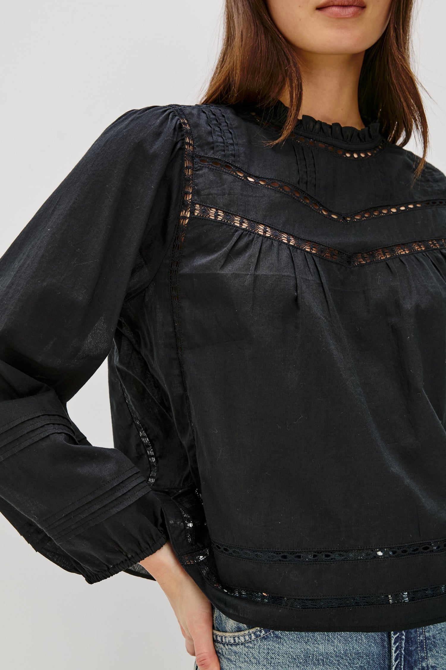 Front detail view of female model wearing our Reyna Top in black. This is a long sleeve crewneck baby doll blouse, with ruffles at the collar. Styled with light wash denim.