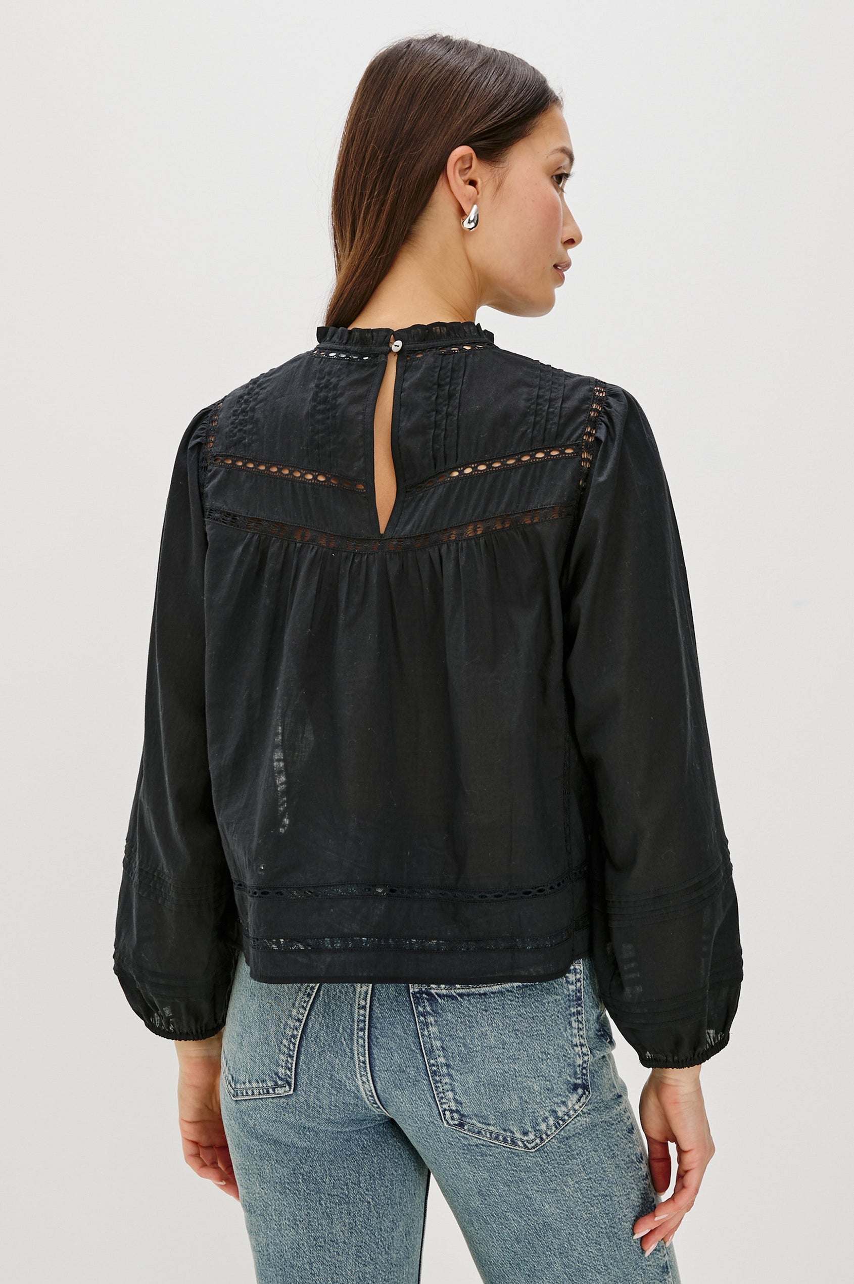 Back view of female model wearing our Reyna Top in black. This is a long sleeve crewneck baby doll blouse, with ruffles at the collar. Styled with light wash denim.