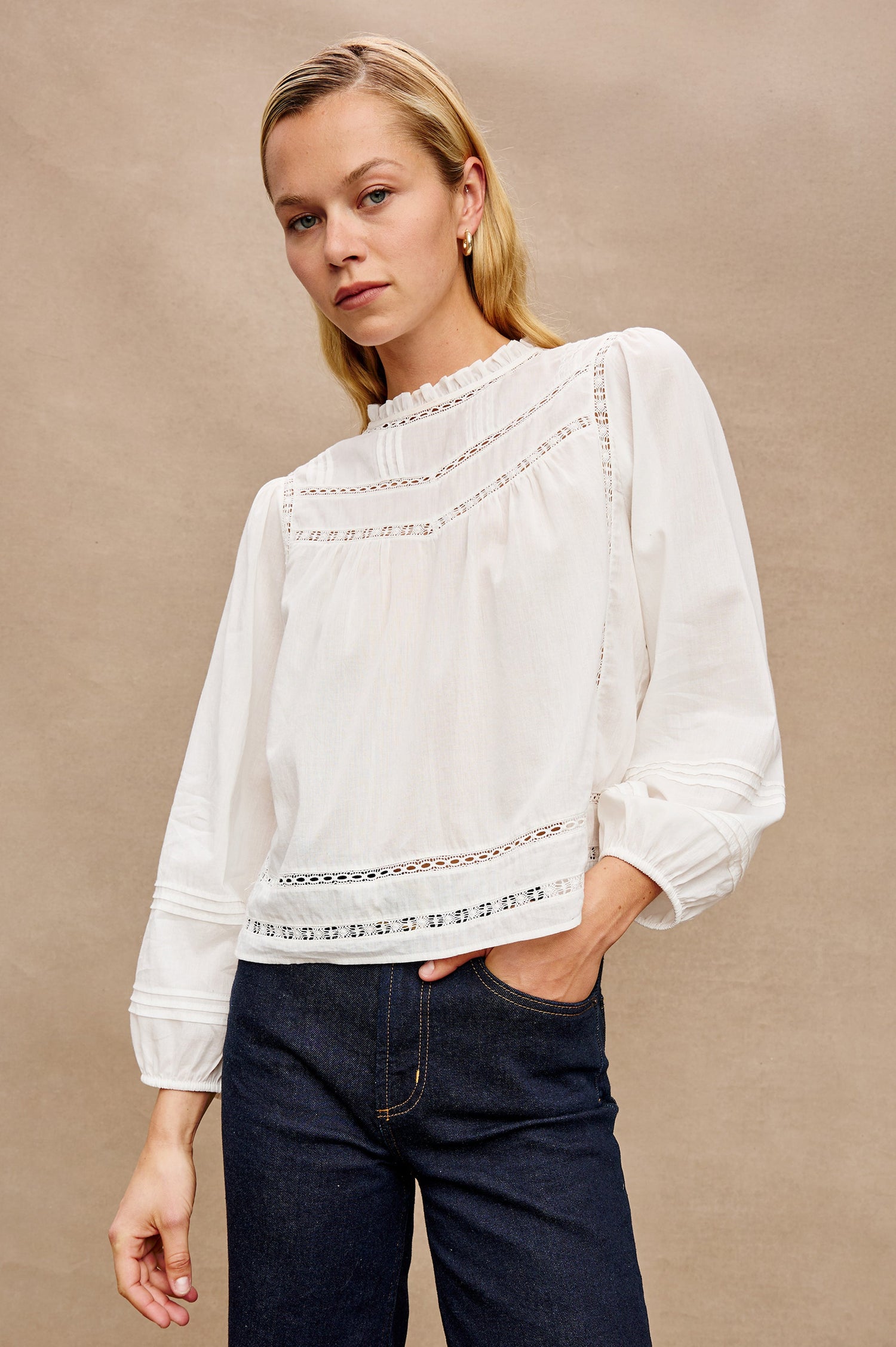 Front view of female model wearing our Reyna Top in white. This is a long sleeve crewneck baby doll blouse, with ruffles at the collar. 
